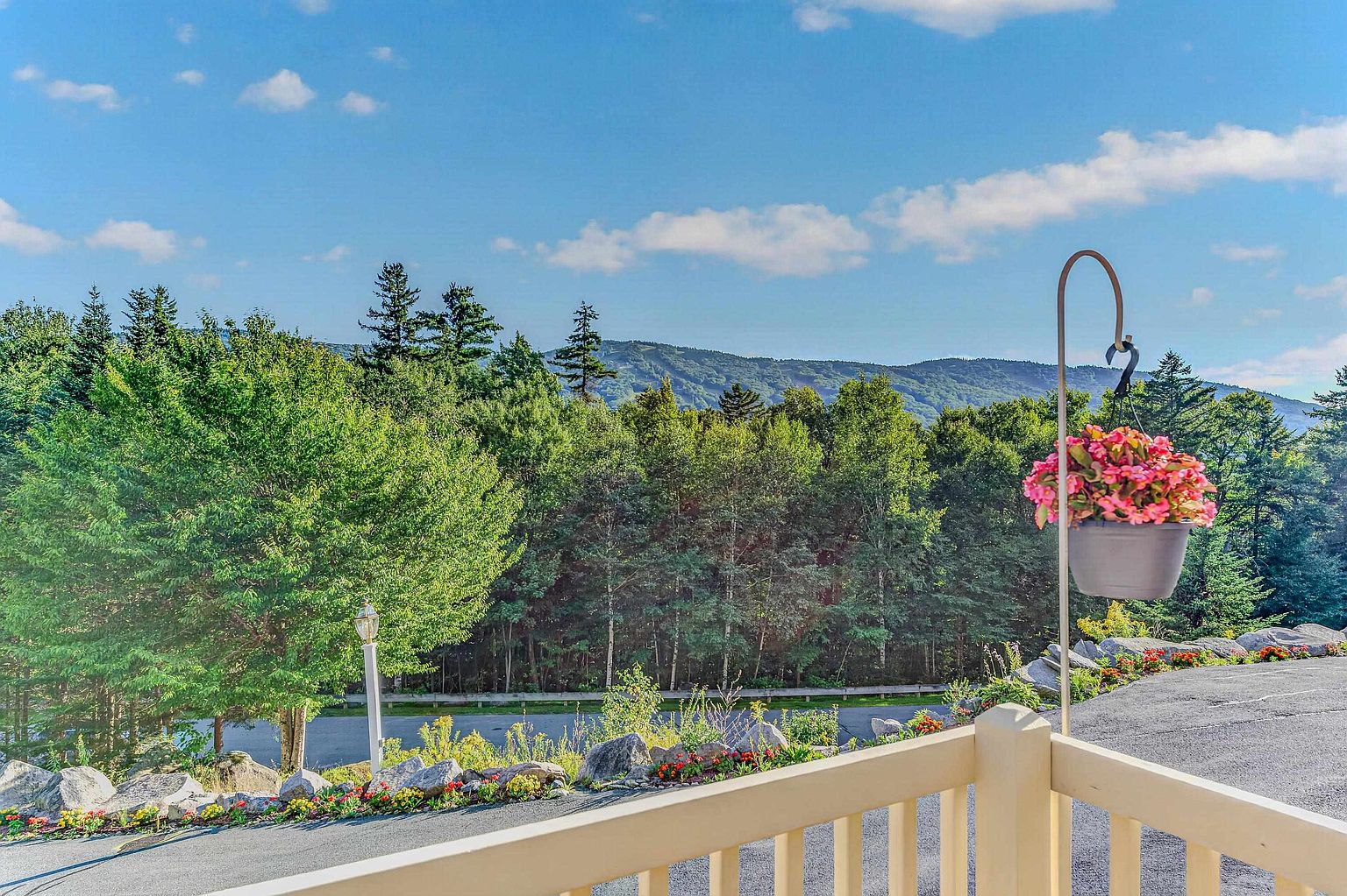 This image captures a scenic view from a balcony, featuring a light-colored railing in the foreground and a hanging basket of vibrant pink flowers. Beyond the railing, a paved driveway leads toward a lush forest and rolling mountains under a bright blue sky with scattered clouds. The perspective offers a peaceful, elevated vantage point that highlights the property's natural surroundings and outdoor living space.