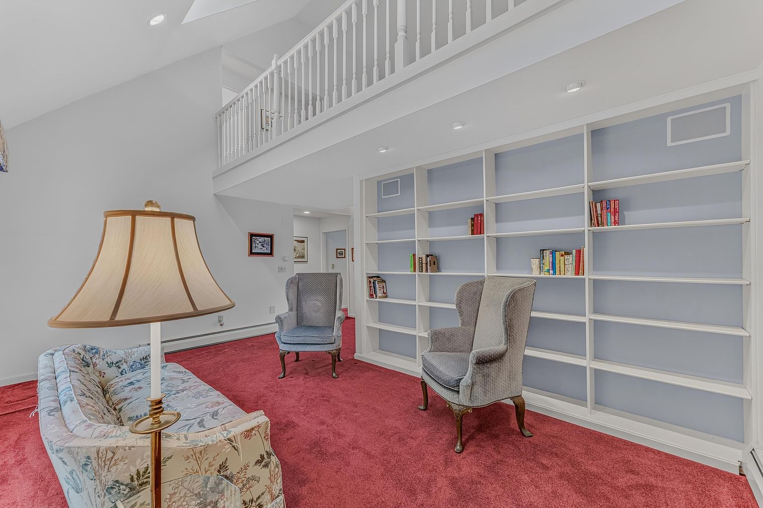 This living room features a vaulted ceiling with a lofted balcony overlooking the space, creating an open and airy atmosphere. The room is furnished with a patterned sofa and two wingback chairs, set against a backdrop of built-in shelving units painted in a soft blue. A prominent floor lamp stands in the foreground, while the red carpeting adds a bold color contrast to the light-toned walls.