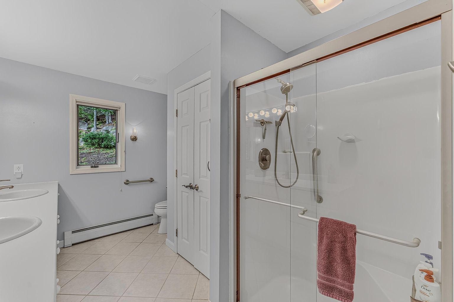 This bright, functional bathroom features a double vanity with light-toned countertops and a walk-in shower enclosure with a glass door and chrome fixtures. A window above the vanity area provides natural light, while a neutral tile floor extends throughout the space. The room includes a toilet tucked into a corner and a white door leading to an adjacent area, creating a clean and accessible layout.