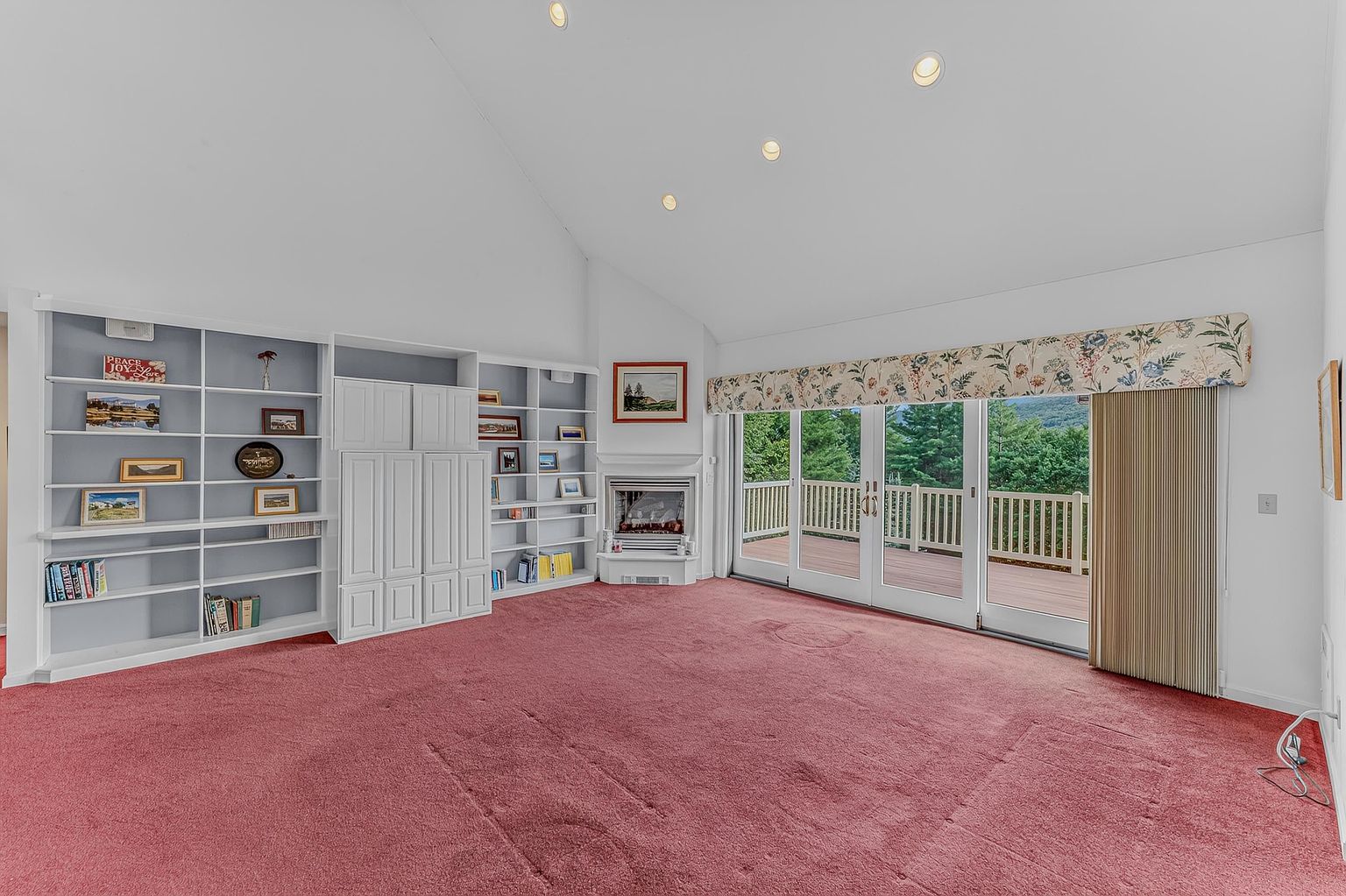 This spacious living room features a vaulted ceiling with recessed lighting and a prominent wall of built-in shelving and cabinetry. A cozy fireplace with a white mantel serves as a focal point, while large sliding glass doors with a floral valance provide access to an outdoor deck and scenic views. The room is carpeted in a deep red, creating a warm and traditional atmosphere.