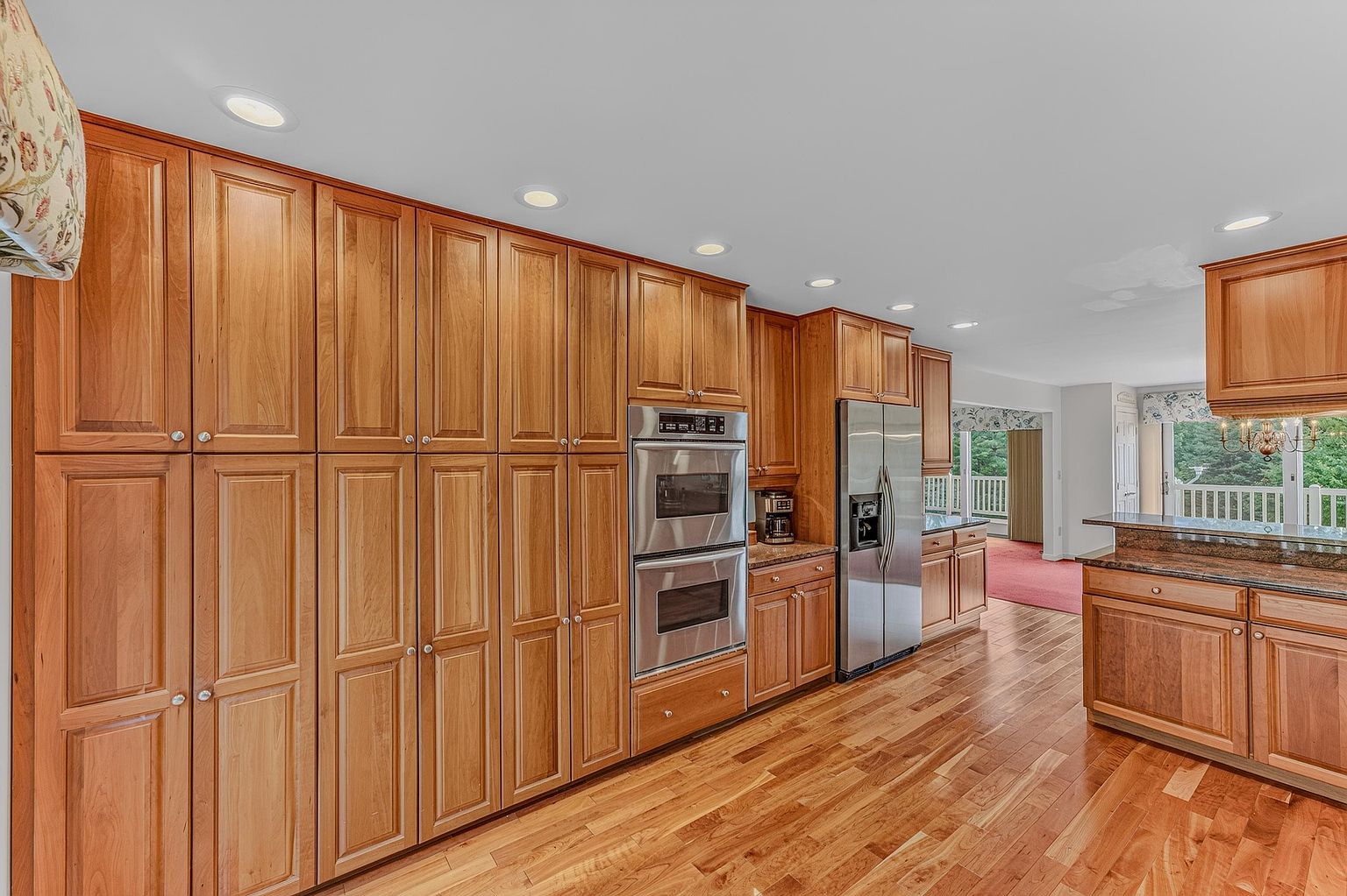 This spacious kitchen features extensive light wood cabinetry and warm-toned hardwood flooring throughout. The layout includes a built-in double oven, a stainless steel refrigerator, and a peninsula with a dark stone countertop that overlooks an adjacent living area. The perspective is a wide-angle shot from the kitchen floor, highlighting the ample storage and the open-concept flow toward the rear deck.