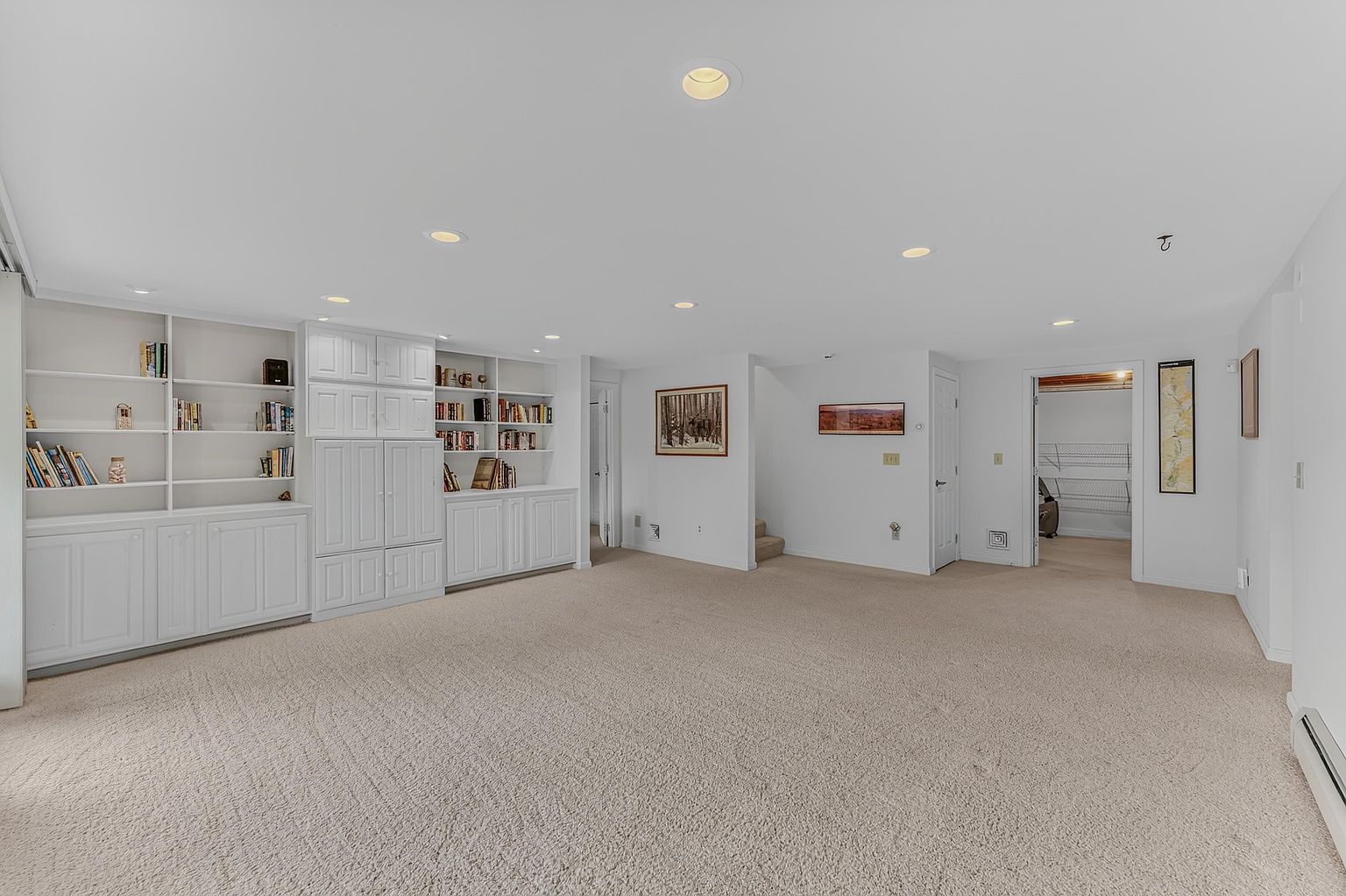 This spacious, carpeted basement features a large built-in white shelving unit and cabinetry along one wall, providing ample storage and display space. The room is brightly lit by recessed ceiling lights and includes a doorway leading to a walk-in storage area, as well as a staircase leading to another level. The neutral color palette and open floor plan create a versatile living space suitable for a recreation room or home office.