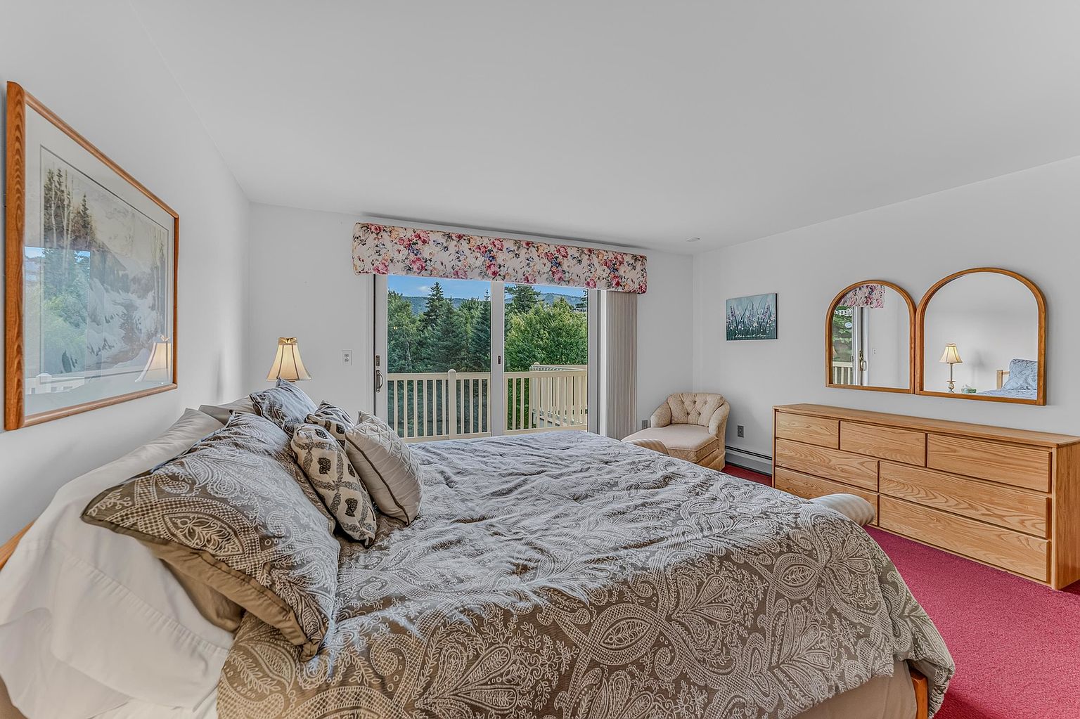 This primary bedroom features a large bed with a patterned comforter as the central focus, positioned in front of a sliding glass door that opens to a balcony with a scenic forest view. To the right, a light wood dresser is topped with two arched mirrors, while a small upholstered chair sits in the corner near the window. The room is finished with red carpeting and a floral valance above the glass door, creating a traditional and cozy atmosphere.