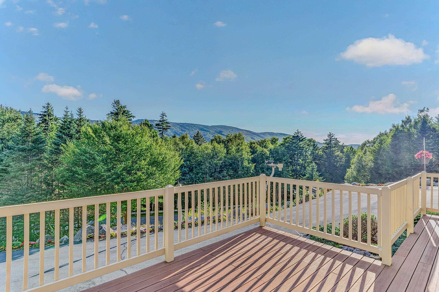 This image captures a spacious, elevated wooden deck featuring light-colored railings and a clear view of a lush, tree-covered mountain range under a bright blue sky. The perspective is from the deck surface, looking out over the railing toward the scenic landscape and a paved area below. The scene conveys a peaceful, open-air atmosphere ideal for outdoor relaxation and enjoying the natural surroundings.