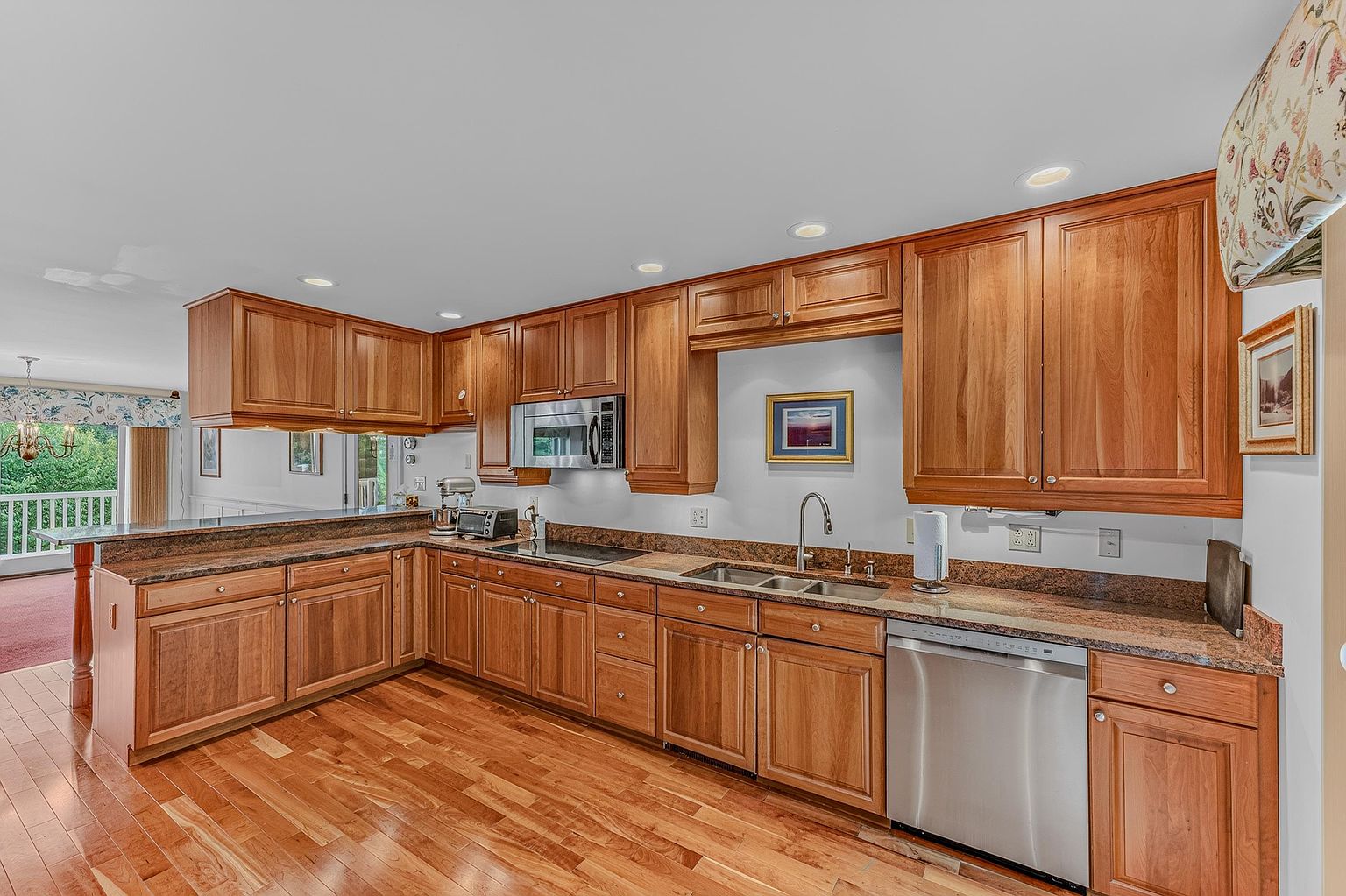 This kitchen features warm-toned wood cabinetry paired with dark stone countertops and a matching backsplash. The space is equipped with stainless steel appliances, including a built-in microwave and a dishwasher, and is illuminated by recessed ceiling lights. A peninsula provides additional counter space and creates an open transition to the adjacent living area, which features a view of an outdoor deck.