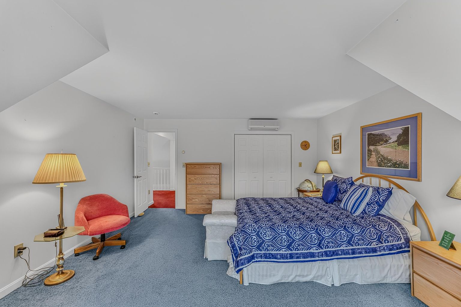 This spacious bedroom features a blue carpeted floor and a large bed with a patterned blue and white duvet cover. The room includes a light wood dresser, a red accent chair, and a bedside lamp, all set against neutral walls under a sloped ceiling. A doorway leads to a hallway, and a wall-mounted air conditioning unit is positioned above the closet doors.
