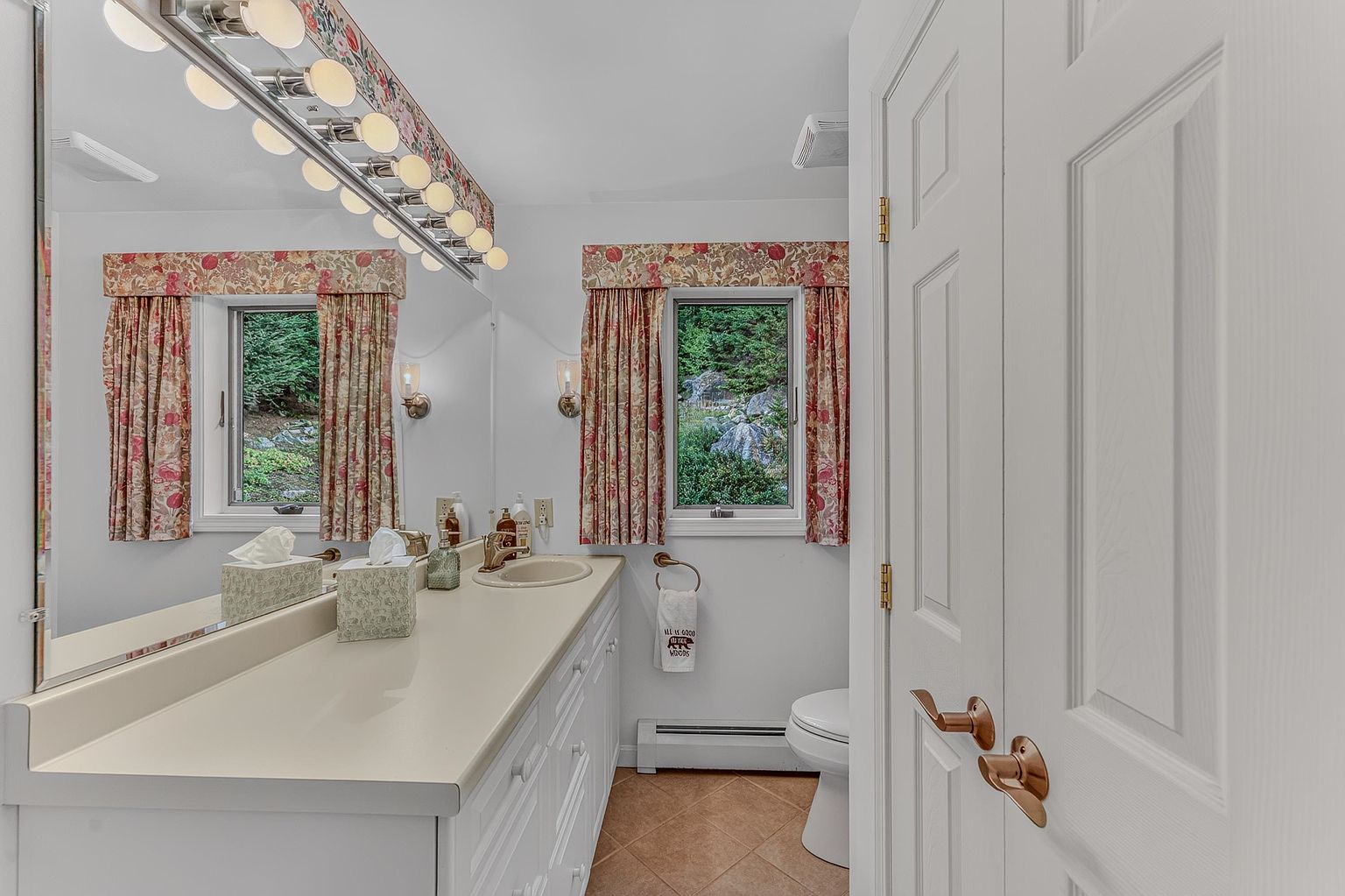 This bright bathroom features a long vanity with a light-colored countertop and white cabinetry, complemented by a large mirror reflecting the space. Floral-patterned valances adorn the windows, which provide a view of the natural, wooded landscape outside. The room is finished with neutral-toned floor tiles and a white toilet, creating a clean and functional atmosphere.