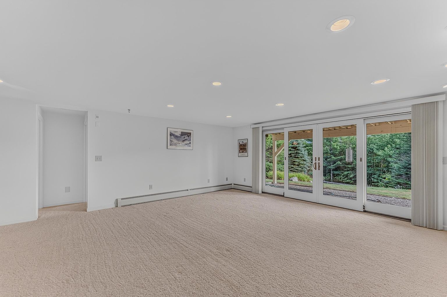 This spacious, neutral-toned living room features wall-to-wall carpeting and a clean, minimalist aesthetic. A large set of sliding glass doors provides abundant natural light and a clear view of the lush, wooded backyard. The room is illuminated by recessed lighting and includes baseboard heating units along the wall.