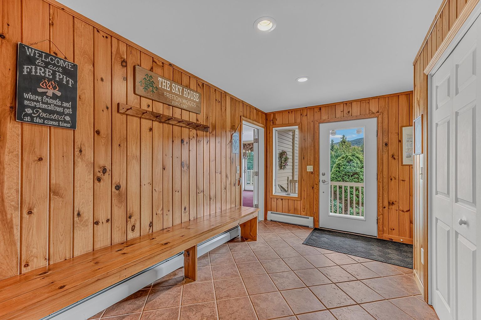 This rustic entryway features warm wood-paneled walls and durable tile flooring, creating a welcoming mountain-lodge aesthetic. A long, built-in wooden bench runs along the wall, complemented by decorative signage and a coat rack, while a glass-paneled door provides a view of the surrounding greenery. The space is illuminated by recessed ceiling lights, offering a clean and functional transition area.