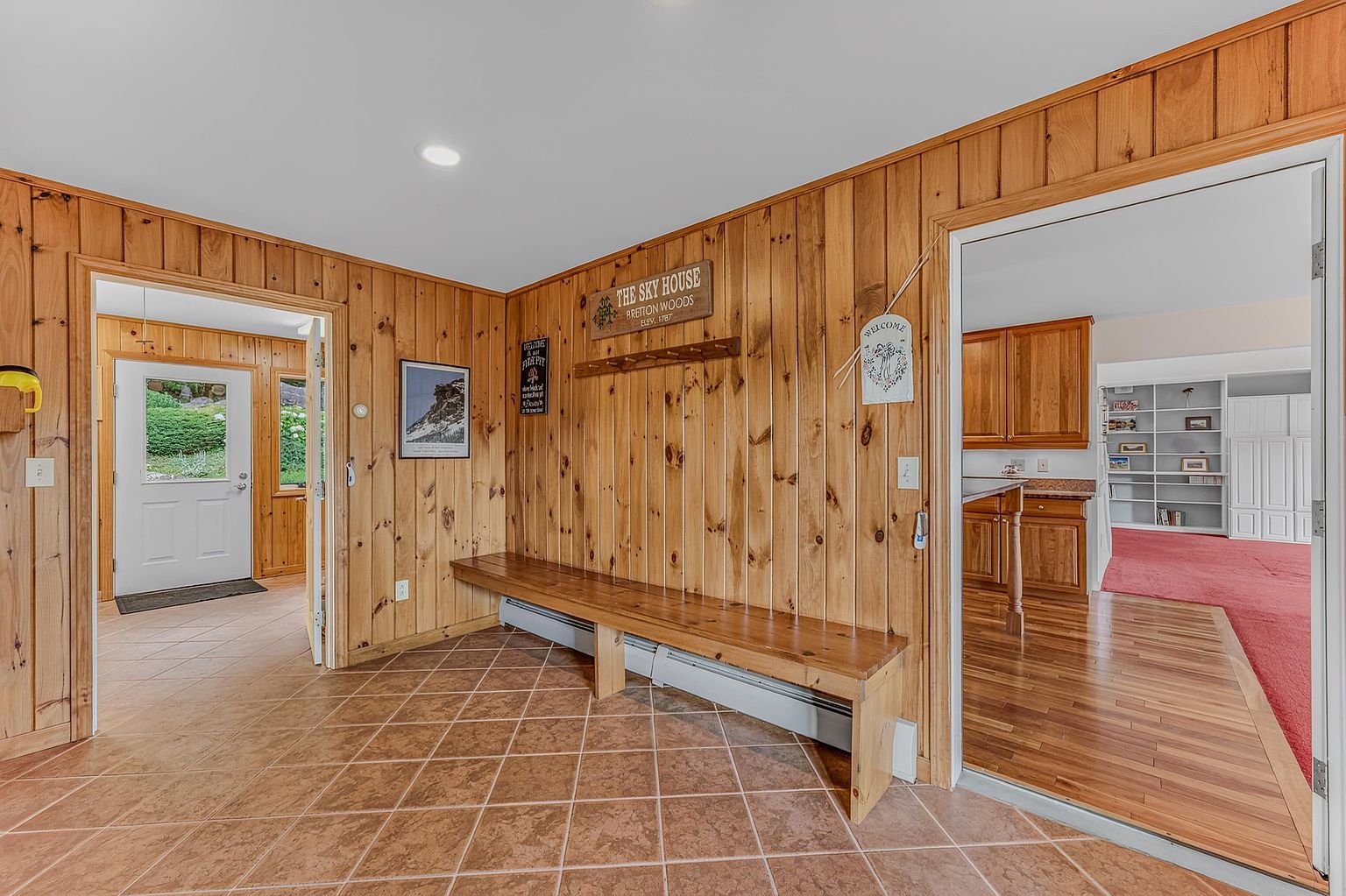 This entryway features warm, vertical wood-paneled walls and a built-in wooden bench, creating a rustic and inviting atmosphere. The floor is covered in large, earth-toned square tiles, and the space provides a clear view into an adjacent kitchen and a carpeted living area. The perspective is centered, showcasing the transition between the tiled foyer and the surrounding living spaces.