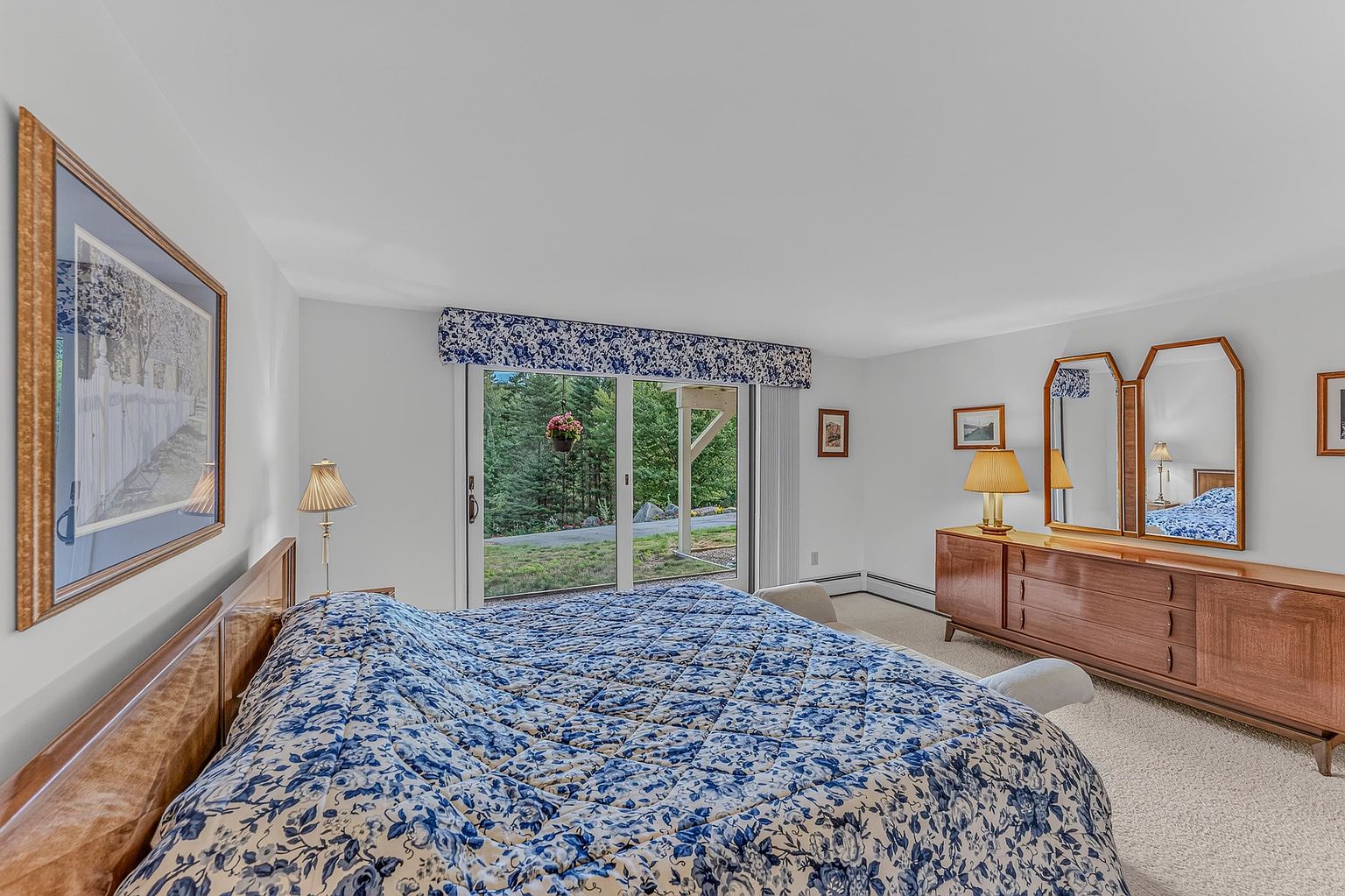 This primary bedroom features a large bed with a blue and white floral quilt, positioned against a light-colored wall. A wooden dresser with a tri-fold mirror sits to the right, complemented by a matching table lamp. Large sliding glass doors provide a view of the wooded exterior, allowing natural light to fill the room.