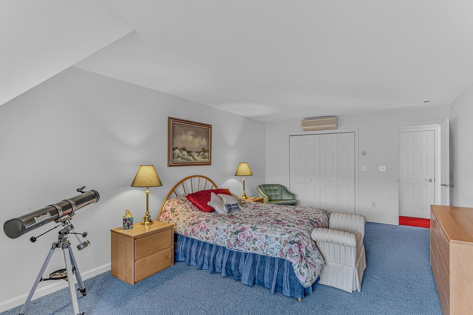 This bedroom features blue carpeting and light-colored walls, centered around a bed with a floral-patterned comforter and a blue bed skirt. A telescope stands in the foreground, while a wooden nightstand with a lamp sits beside the bed, complemented by a framed landscape painting on the wall. The room includes a closet with white doors, a small green armchair, and a light wood dresser, creating a functional and traditional living space.