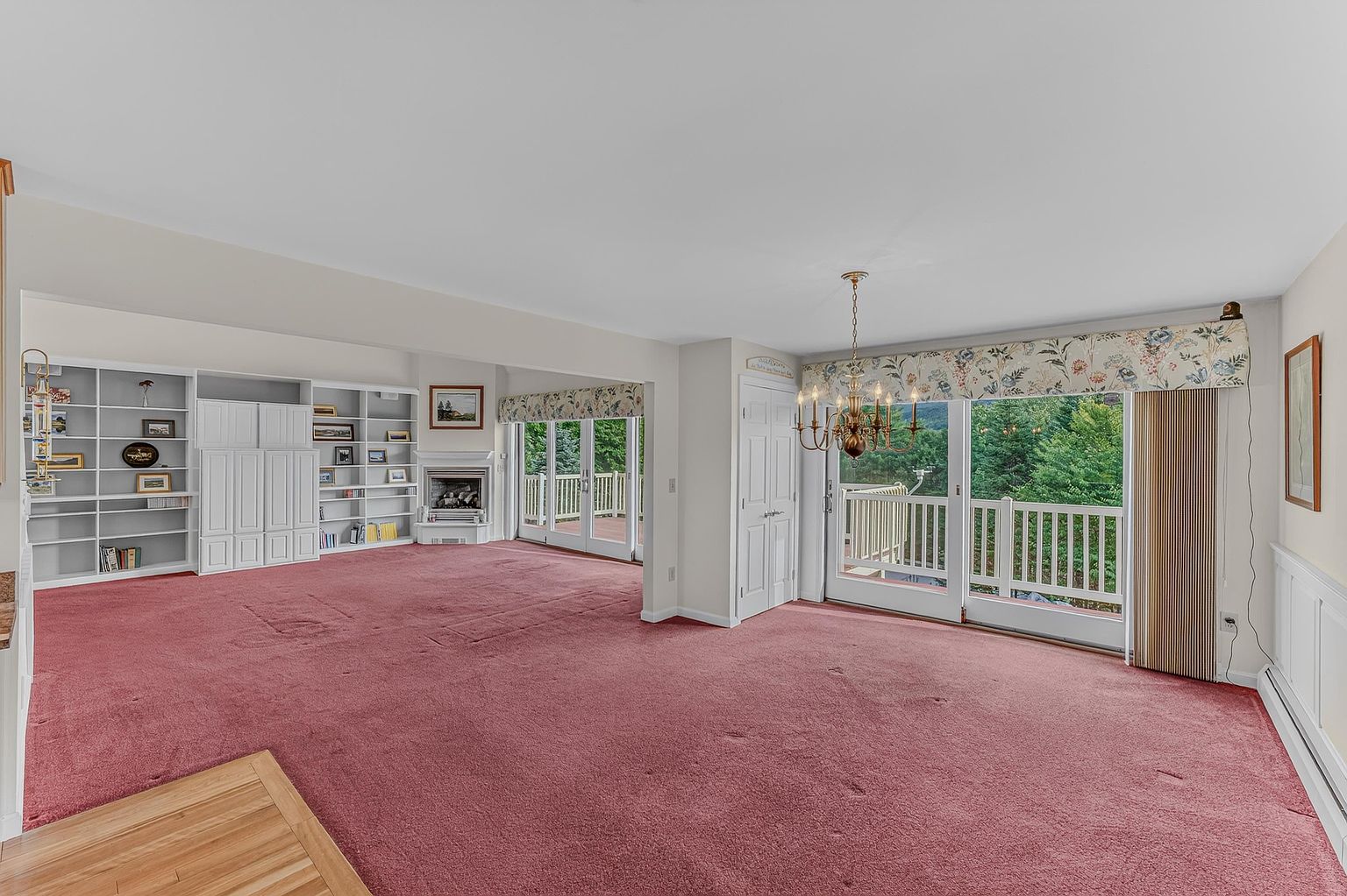 This spacious living room features wall-to-wall carpeting and an expansive built-in shelving unit that provides ample storage and display space. A white fireplace with a mantel serves as a focal point, while large glass sliding doors lead out to a deck, allowing for plenty of natural light. The room is illuminated by a traditional chandelier and includes a decorative floral valance above the glass doors, creating a bright and inviting atmosphere.