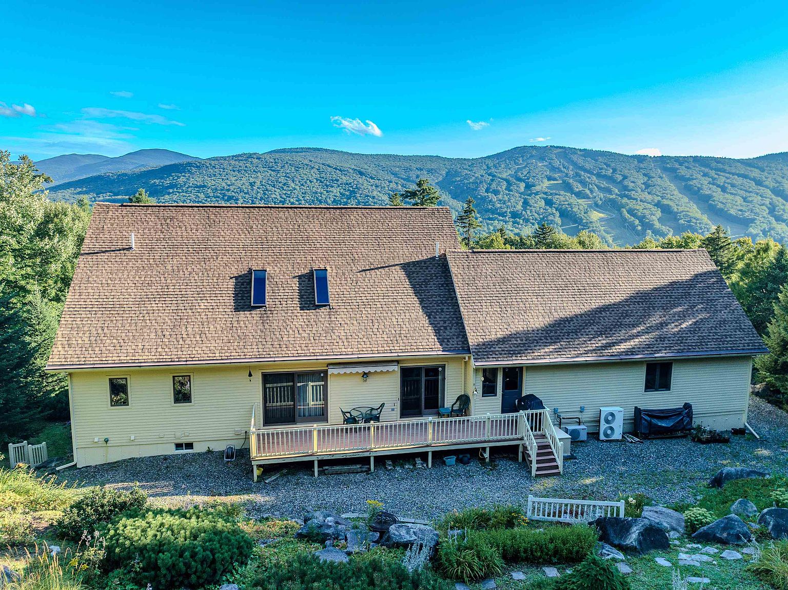 This elevated rear view captures a spacious, light-yellow sided home nestled against a backdrop of lush, rolling mountains. The property features a wide wooden deck spanning the back of the house, providing an ideal space for outdoor relaxation, while the foreground showcases a landscaped garden with stone accents and a gravel yard. The scene is bathed in bright, clear daylight, emphasizing the home's scenic location and peaceful atmosphere.
