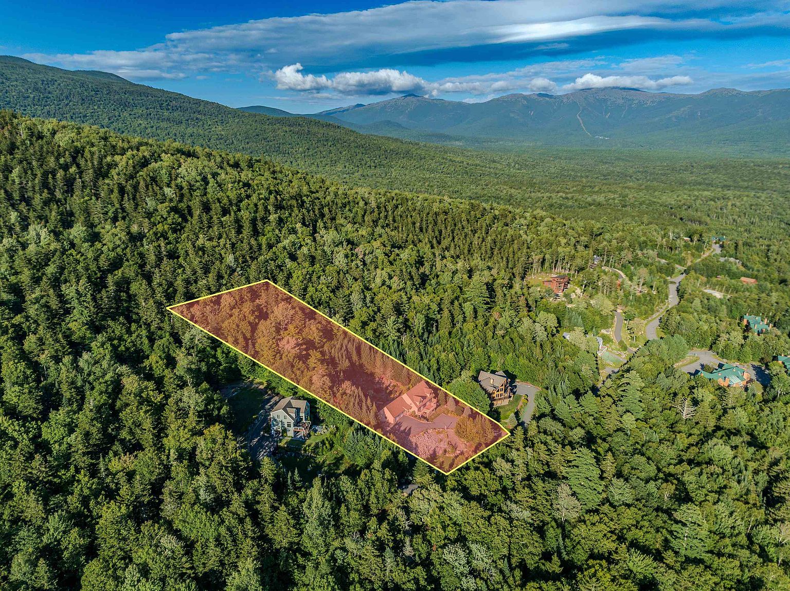 This high-angle aerial view captures a residential property nestled within a dense, lush forest, with a highlighted lot boundary overlaying the wooded landscape. The scene showcases a multi-story home with a driveway and surrounding trees, set against a backdrop of rolling mountain ranges under a clear blue sky. The perspective emphasizes the secluded, nature-oriented setting of the home within a larger mountainous region.
