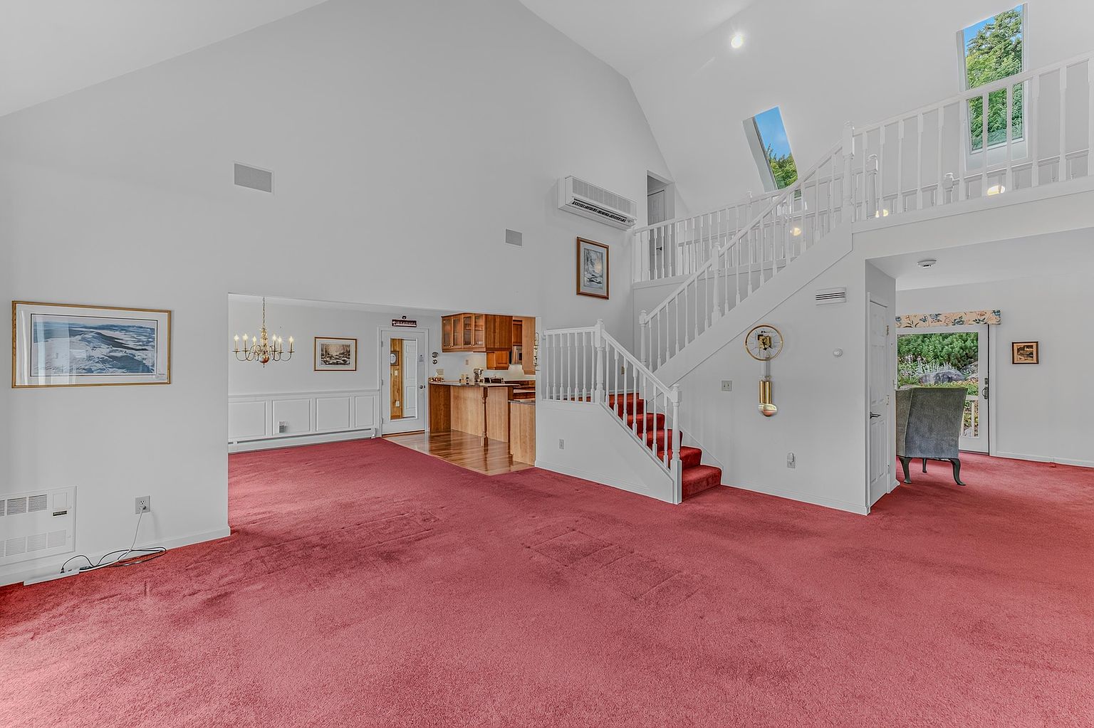 This spacious, open-concept living area features a dramatic vaulted ceiling and a prominent staircase with a white railing leading to an upper landing. The room is carpeted in a bold red, creating a striking contrast against the neutral white walls, while a glimpse into the adjacent kitchen reveals warm-toned wood cabinetry. A large window and a skylight allow natural light to fill the space, which is accented by a traditional grandfather clock and framed wall art.