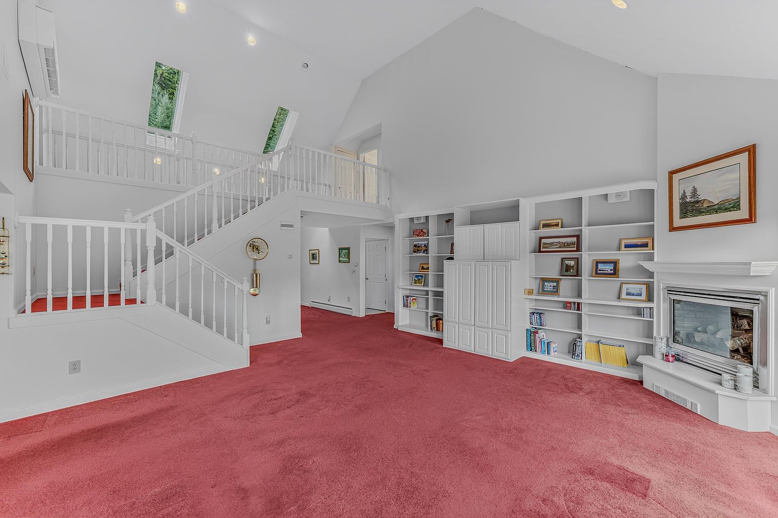This spacious living room features a vaulted ceiling and a prominent staircase with white railings leading to an upper-level landing. The room is carpeted in a bold red and includes a built-in white shelving unit flanking a fireplace with a mantel and a framed painting above. The open-concept design creates an airy atmosphere, with natural light filtering in from high-placed windows.