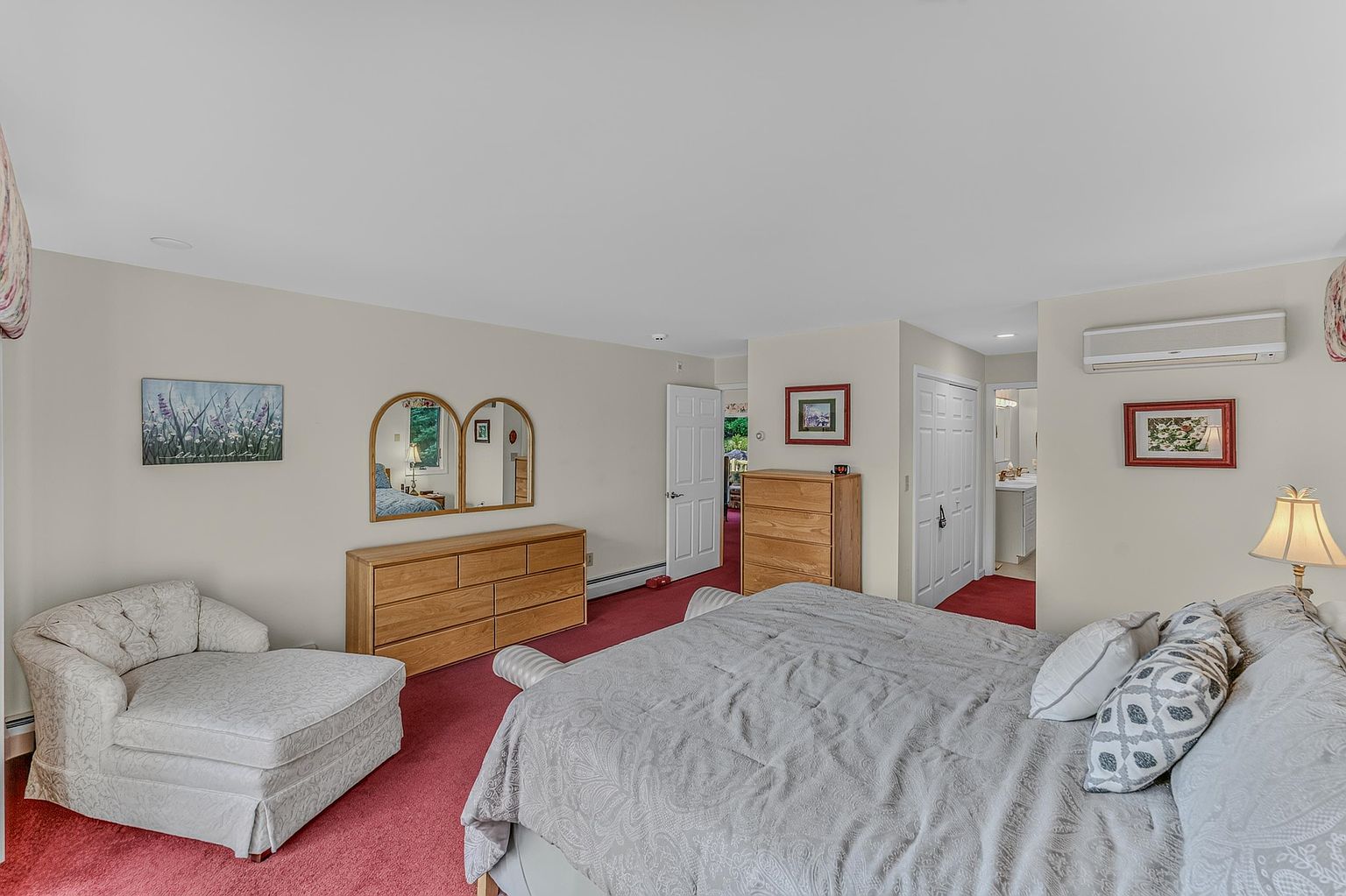 This spacious primary bedroom features a neutral color palette, red carpeting, and a large bed with patterned bedding. The room includes a light wood dresser with a mirror, a comfortable upholstered chaise lounge, and a wall-mounted air conditioning unit. A doorway leads to an adjacent bathroom, and the space is accented with framed wall art.
