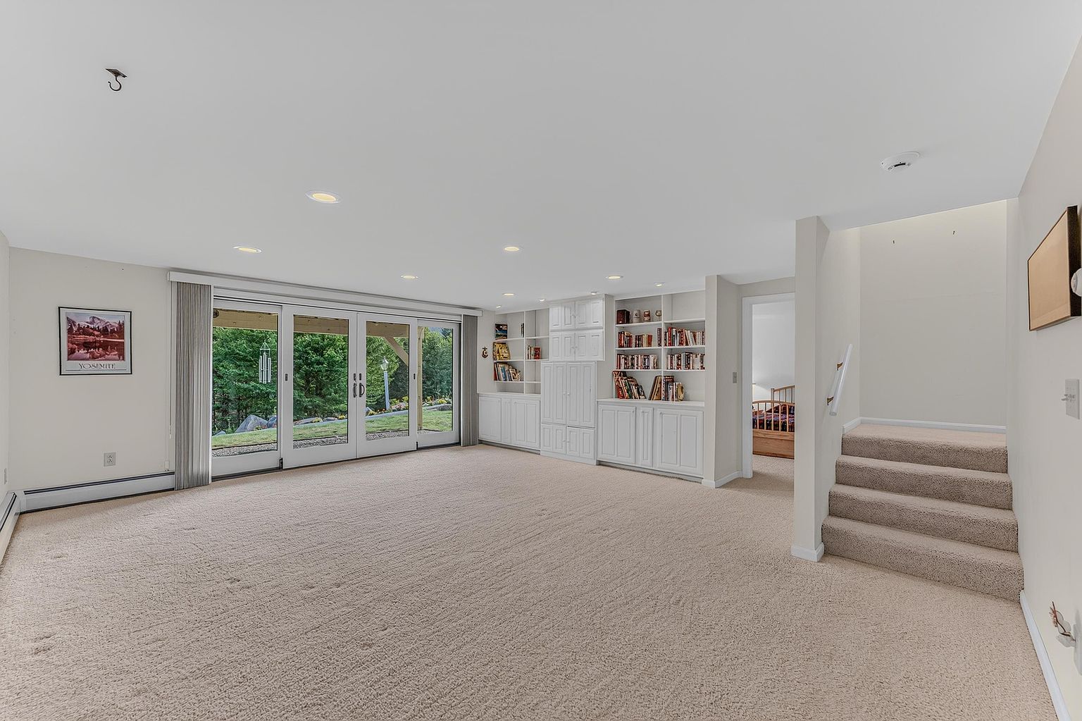 This spacious, carpeted basement features a large wall of glass doors that provide abundant natural light and a view of the outdoor greenery. The room includes built-in white cabinetry and shelving units, offering ample storage and display space. A set of carpeted stairs leads to an upper level, while a doorway reveals a glimpse into an adjacent bedroom, creating an open and versatile living area.