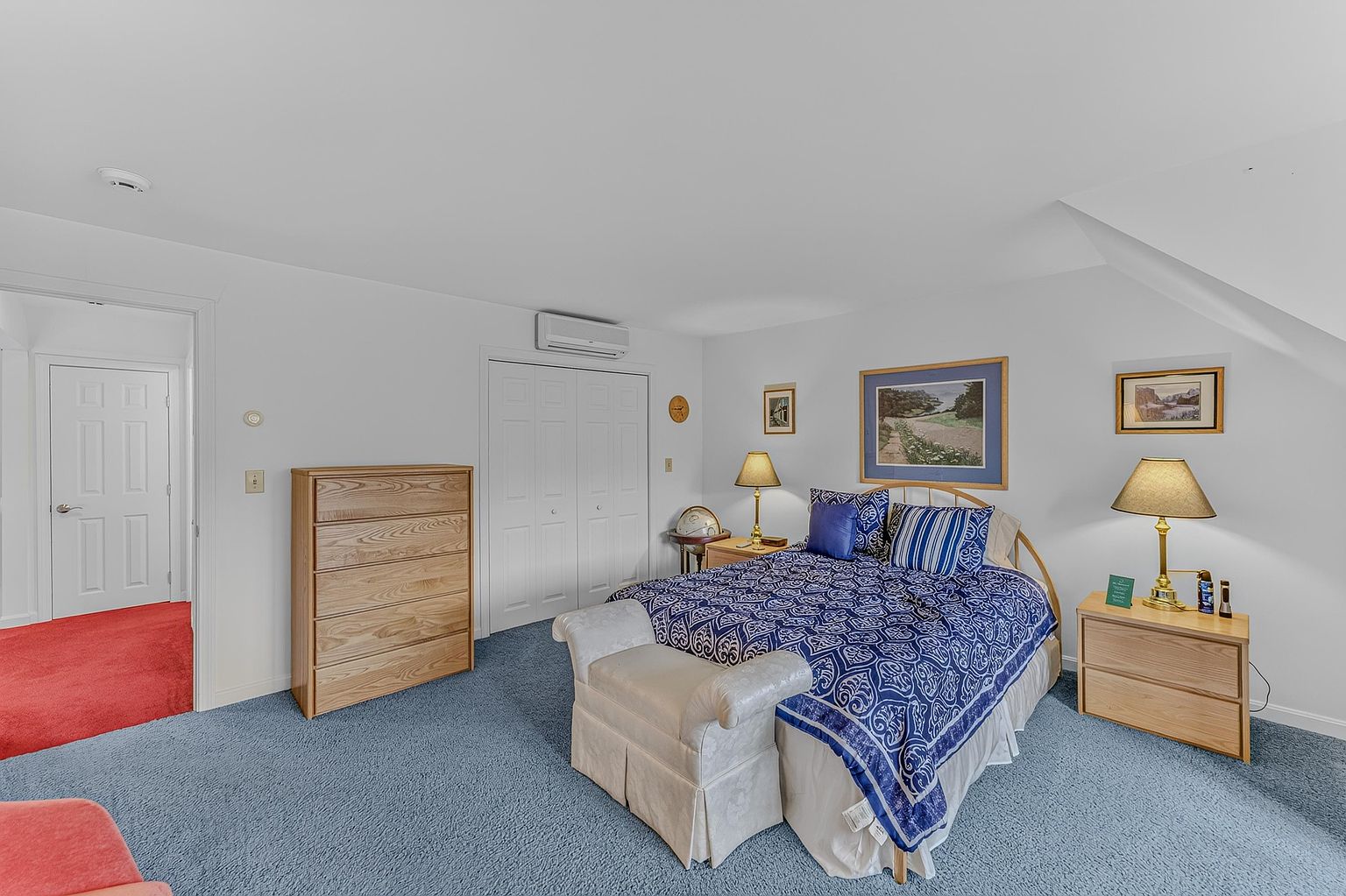 This bedroom features a queen-sized bed with a bold blue and white patterned comforter, flanked by two light wood nightstands with matching lamps. A light wood dresser sits against the wall, and a white upholstered bench is positioned at the foot of the bed. The room is carpeted in blue and includes a white closet with double doors and a wall-mounted air conditioning unit.