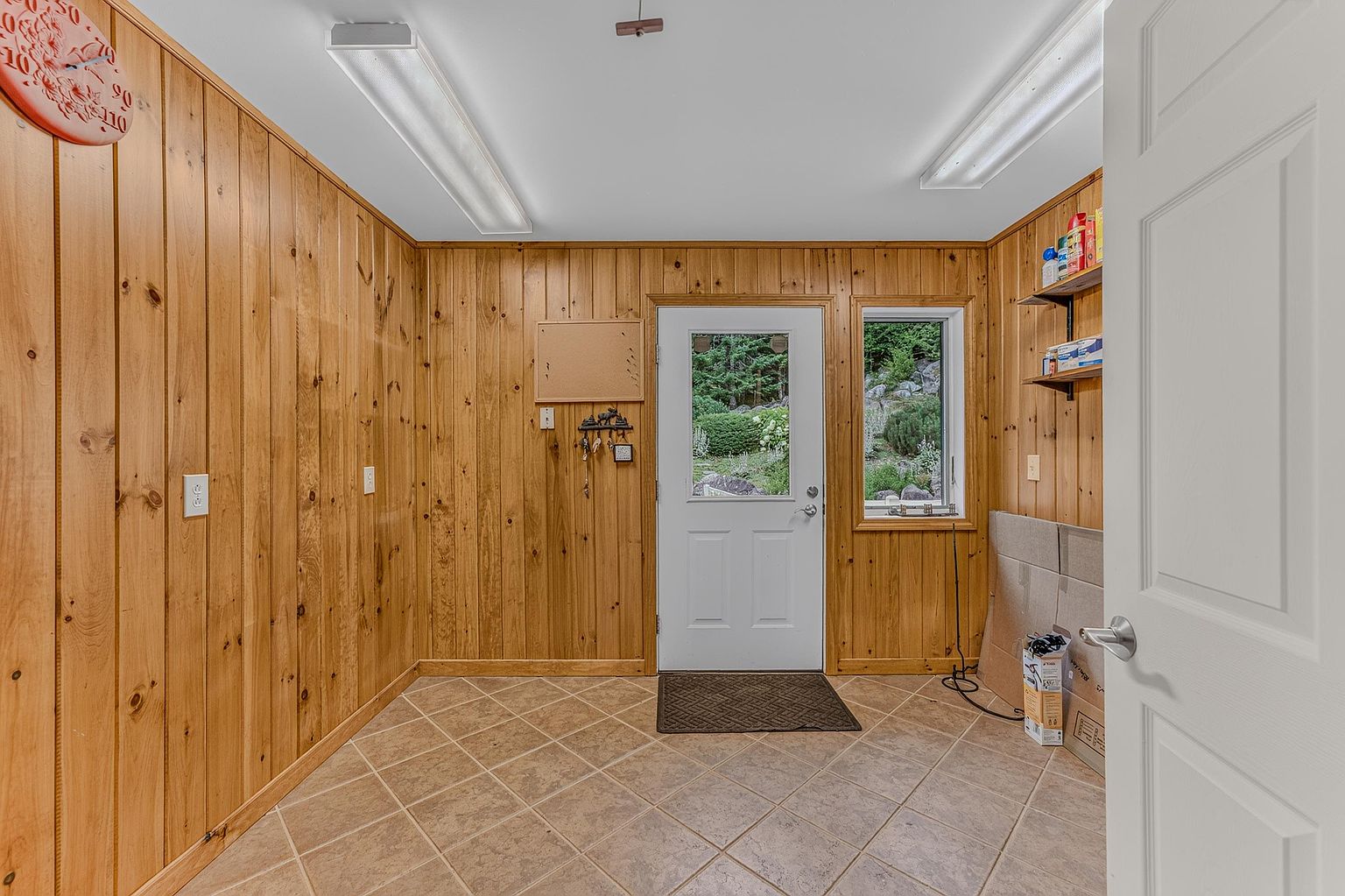 This entryway features warm, wood-paneled walls and neutral-toned tile flooring, creating a rustic and functional space. A central white door with a glass insert provides natural light and a view of the outdoor greenery, complemented by a nearby window. The room is illuminated by two long, rectangular ceiling-mounted light fixtures and includes practical storage elements like wall-mounted hooks and open shelving.