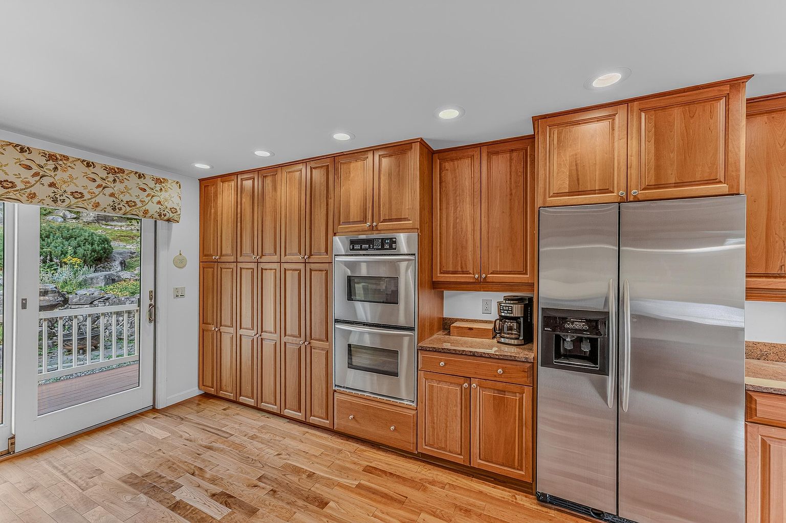 This kitchen features warm-toned wood cabinetry, stainless steel appliances, and light-colored stone countertops. A double wall oven is integrated into the cabinetry, and a large stainless steel refrigerator stands to the right. A glass door with a patterned valance provides a view of the outdoor deck and greenery, while recessed lighting illuminates the hardwood floors.