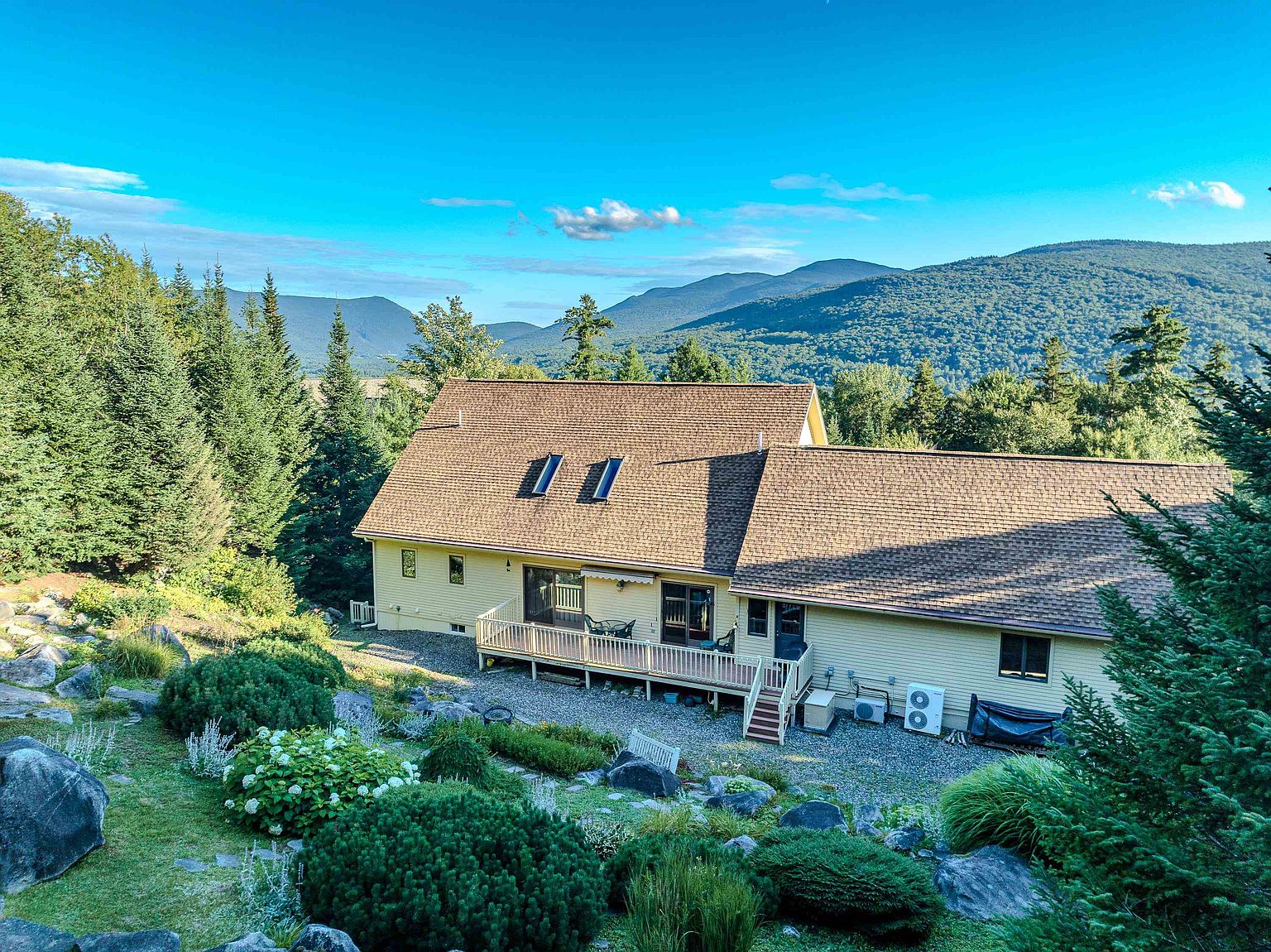 This elevated perspective captures the rear of a light-yellow, multi-level home nestled against a backdrop of lush, rolling mountains. The property features a spacious wooden deck spanning the back of the house, complemented by a landscaped garden filled with shrubs, boulders, and perennial plantings. The scene conveys a serene, private retreat with a clear view of the expansive natural landscape.