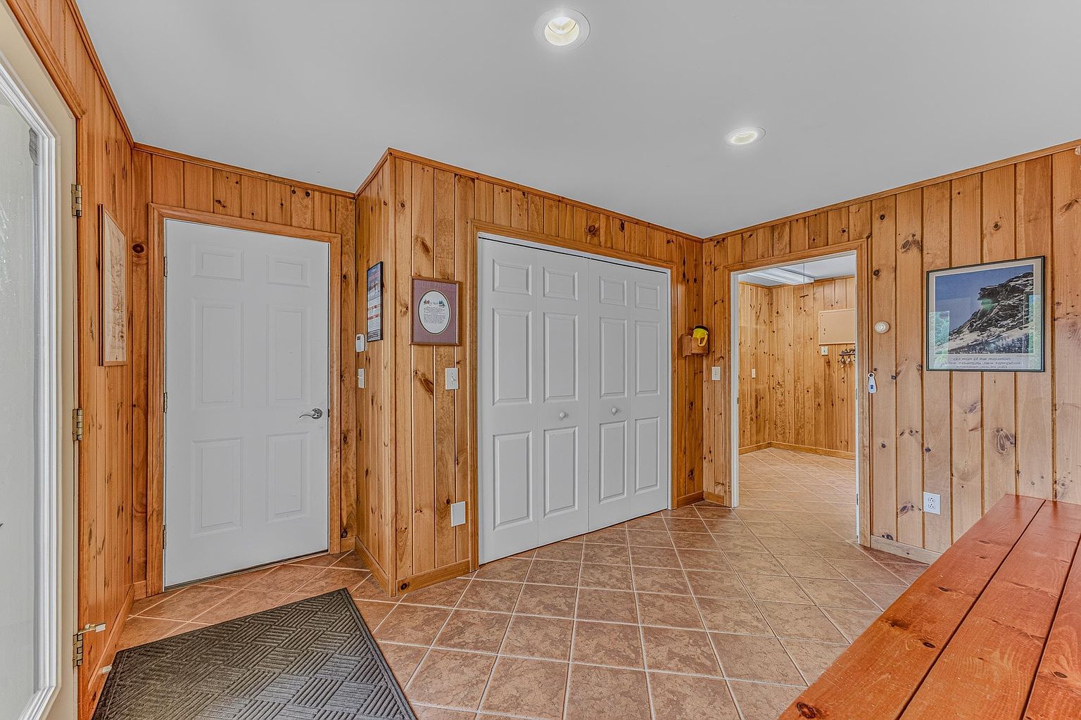 This entryway features warm wood-paneled walls and neutral-toned tile flooring, creating a rustic and inviting atmosphere. A white door provides access to the exterior, while a set of double closet doors offers convenient storage space. The perspective looks into an adjacent room, showcasing the consistent wood paneling throughout the home's interior.