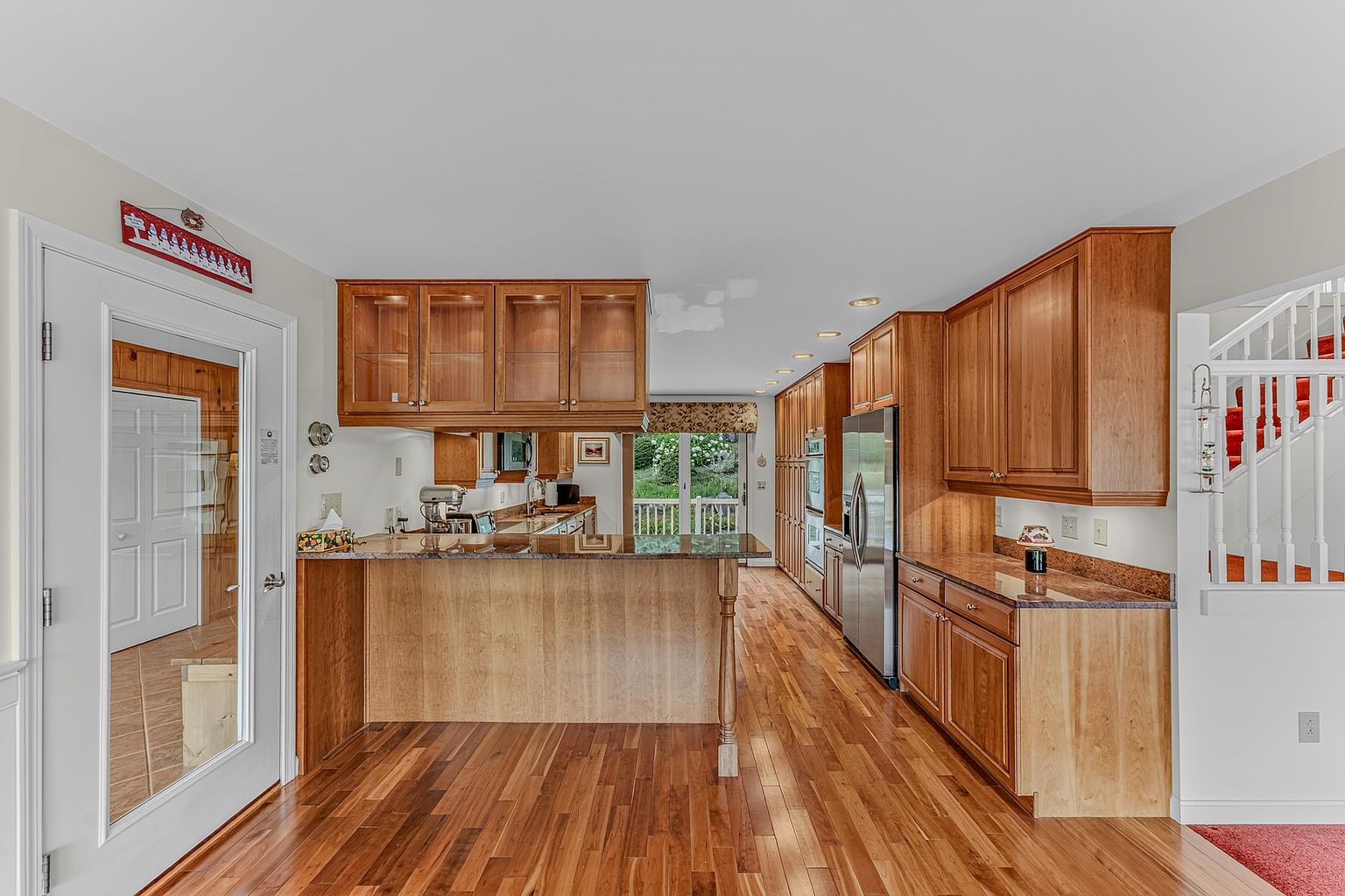 This spacious kitchen features warm-toned wood cabinetry, a large peninsula with a stone countertop, and hardwood flooring throughout. The layout includes a stainless steel refrigerator, built-in wall ovens, and glass-front upper cabinets, all illuminated by recessed lighting. A glass door on the left provides a view into an adjacent room, while a set of French doors at the far end leads to an outdoor space, creating an open and inviting atmosphere.
