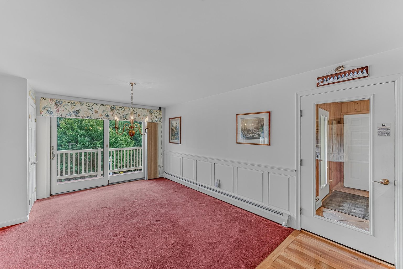 This interior space features a neutral color palette with white walls and wainscoting, contrasted by a bold red carpet. A large sliding glass door with a floral valance provides access to an outdoor balcony, while a classic chandelier hangs centrally in the room. The perspective captures the transition from the carpeted living area to a wood-floored entryway, showcasing a bright and traditional layout.