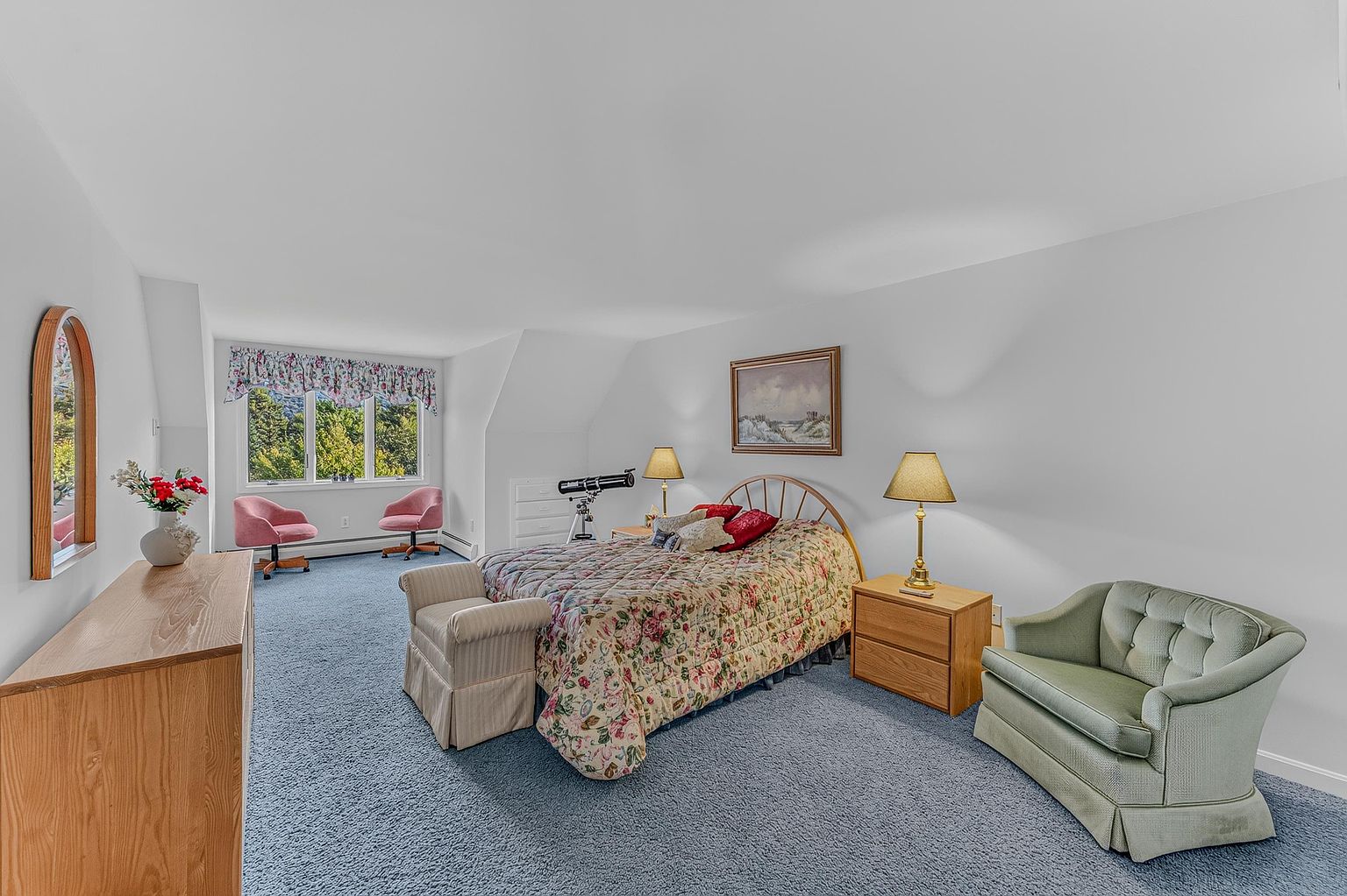 This spacious bedroom features a floral-patterned bedspread, a light wood dresser, and a matching nightstand with a lamp. A soft green armchair sits in the corner, while two pink chairs are positioned near the large windows that offer a view of the outdoors. The room is carpeted in a textured blue, creating a traditional and cozy atmosphere.
