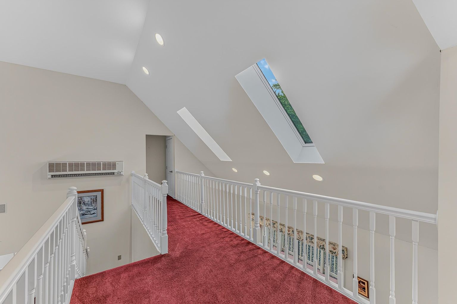 This interior view captures a carpeted upper-level hallway or landing area, characterized by a dramatic vaulted ceiling and white railings. Two skylights are integrated into the sloped ceiling, allowing natural light to brighten the space, while recessed lighting provides additional illumination. The perspective looks down the length of the walkway, highlighting the open, airy feel of the architectural design.