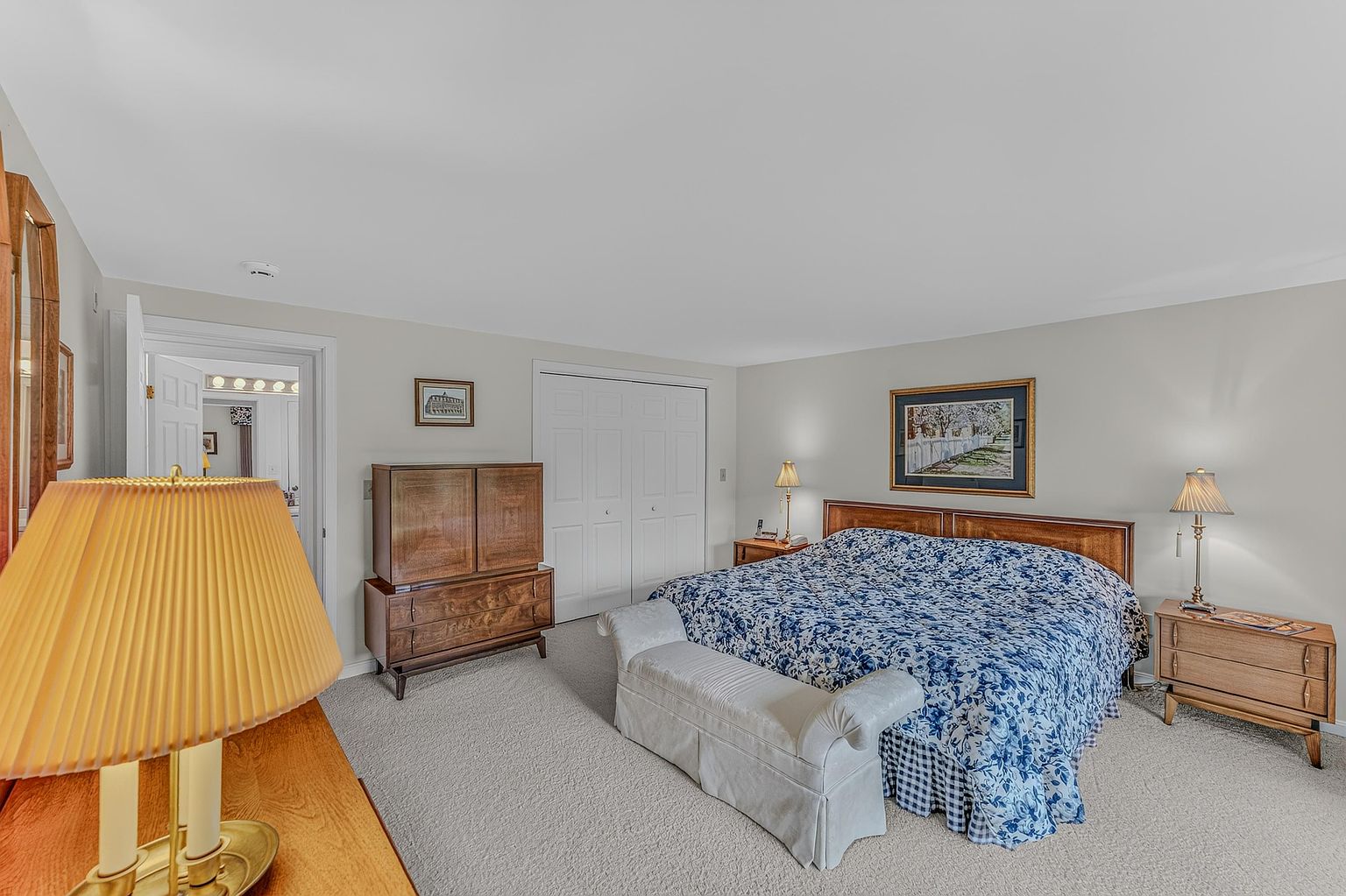 This spacious primary bedroom features a large bed with a blue and white floral patterned comforter, flanked by two matching wooden nightstands with lamps. A light-colored upholstered bench sits at the foot of the bed, while a tall wooden armoire provides additional storage against the wall. The room is carpeted in a neutral tone and includes a doorway leading to an adjacent bathroom, creating a comfortable and traditional living space.