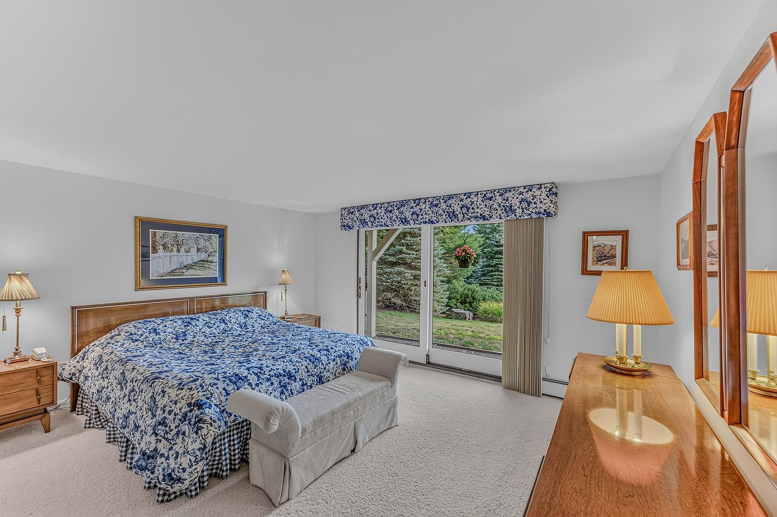 This spacious primary bedroom features a large bed with a blue and white floral patterned comforter and a matching valance above the sliding glass doors. The room is furnished with a wooden headboard, matching nightstands with lamps, and a light-colored upholstered bench at the foot of the bed. Large sliding glass doors provide a clear view of the lush green backyard, filling the room with natural light.
