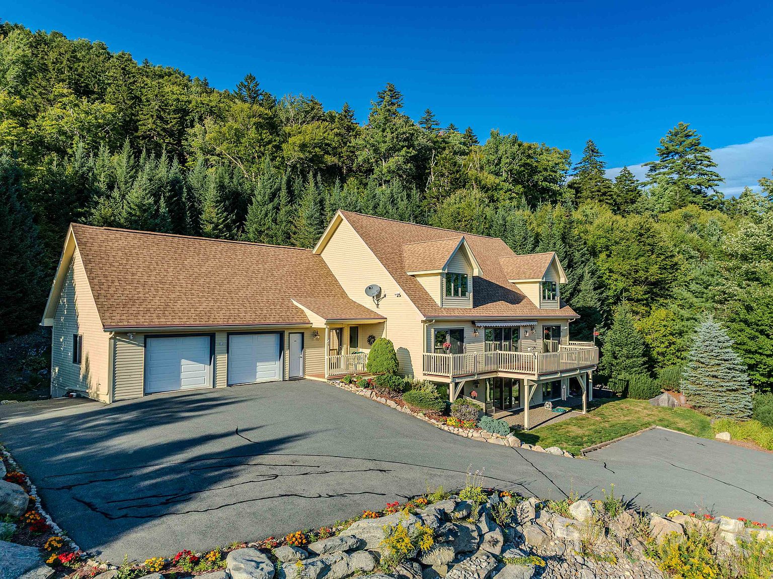 This elevated, wide-angle shot captures a two-story suburban home nestled against a lush, forested hillside. The house features light-colored siding, a multi-gabled roof, and an attached two-car garage, complemented by a spacious wooden deck that overlooks a paved driveway and a landscaped rock garden in the foreground. The bright, clear sky and dense surrounding greenery create a serene and inviting atmosphere for this well-maintained property.