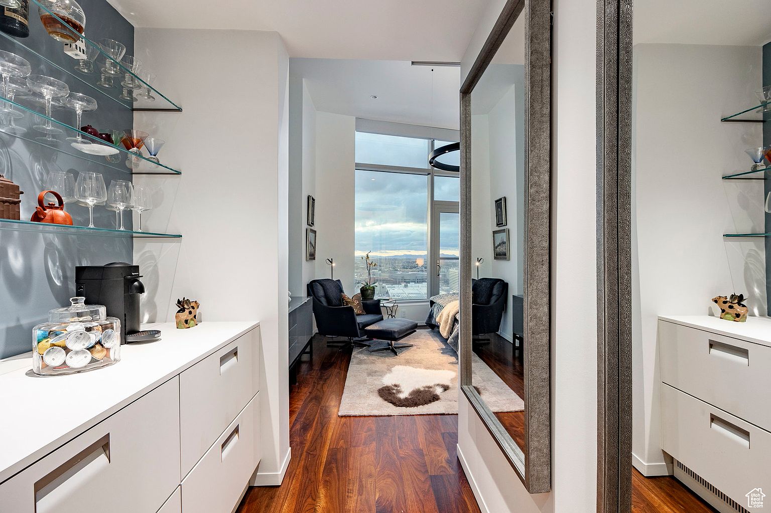 This interior shot showcases a modern hallway leading into a living area with a large window offering city views. The hallway features built-in white cabinetry with glass shelves displaying glassware, a coffee machine, and decorative items. A large mirror reflects the living area, creating a sense of spaciousness, while the hardwood flooring adds warmth to the contemporary design.