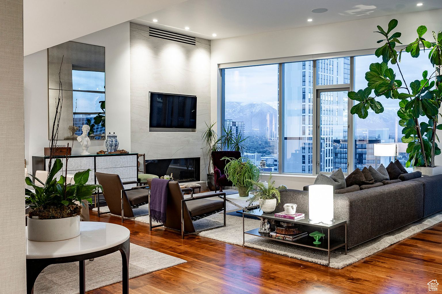 This is a well-lit living room featuring hardwood floors, a modern fireplace with a mounted TV, and large windows offering city views. The room is furnished with a gray sofa, two armchairs, and a coffee table, complemented by indoor plants that add a touch of nature. The overall impression is one of contemporary elegance and comfort.