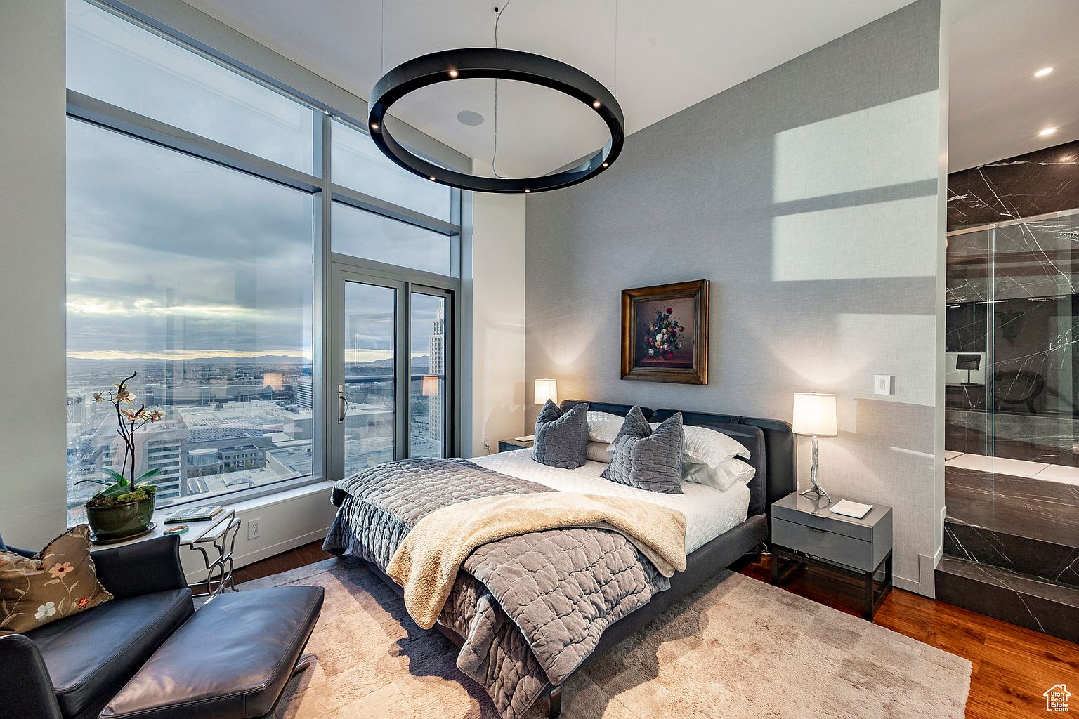 This is a luxurious primary bedroom featuring a large window with a city view, a modern circular light fixture, and a gray textured wall. The bed is adorned with a gray quilted comforter and plush pillows, complemented by stylish nightstands and lamps. The room exudes a sophisticated and comfortable ambiance.