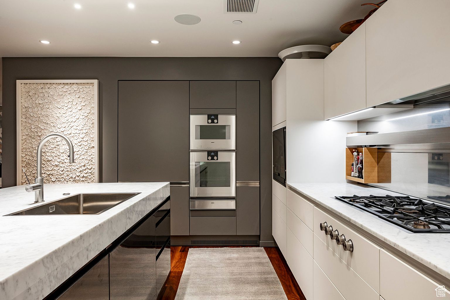 This is a modern kitchen with a sleek design. It features a large island with a marble countertop and a stainless steel sink. The kitchen also has gray and white cabinets, stainless steel appliances, and a hardwood floor with a rug. The perspective is from the front, showcasing the island and the appliances.