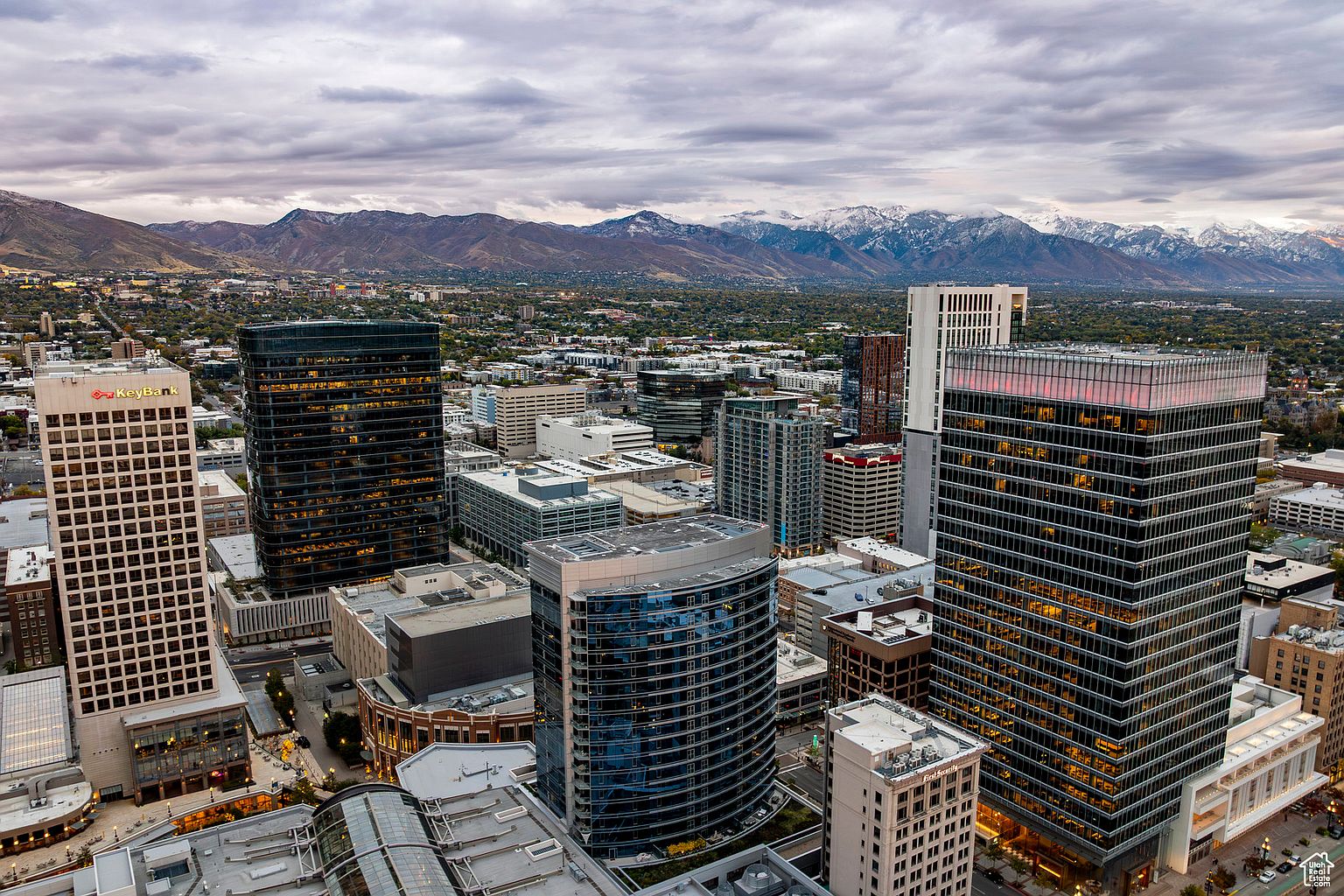 This aerial view showcases a dense urban landscape with numerous high-rise buildings, set against a backdrop of distant mountains with snow-capped peaks. The city features a mix of modern and classic architectural styles, with a variety of building heights and designs. The overall impression is one of a thriving metropolitan area nestled within a stunning natural environment.