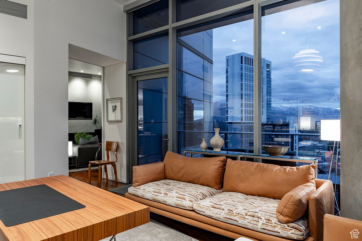 This is an interior shot of a modern living room featuring a brown leather sofa with patterned cushions, a wooden desk with a black top, and floor-to-ceiling windows offering a city view. The room is well-lit, and the decor is minimalist, creating a sophisticated and inviting atmosphere. The perspective is from a medium shot, showcasing the room's layout and design elements.