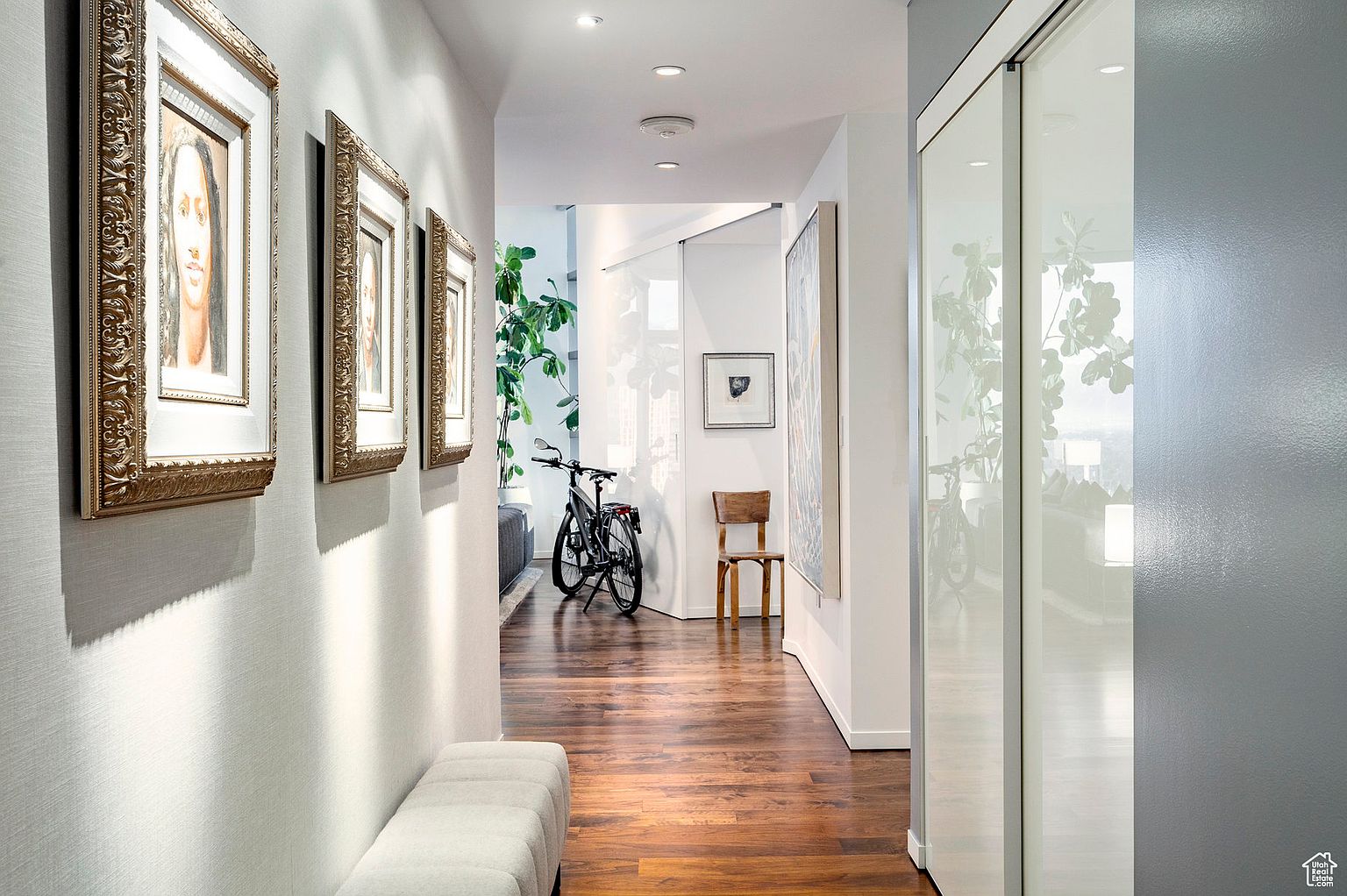 This is an interior shot of a well-lit hallway featuring hardwood floors and white walls adorned with framed artwork. A bench sits along the wall, and a bicycle is parked further down the hall, adding a touch of modern living. The perspective draws the eye down the hallway, creating a sense of depth and spaciousness.