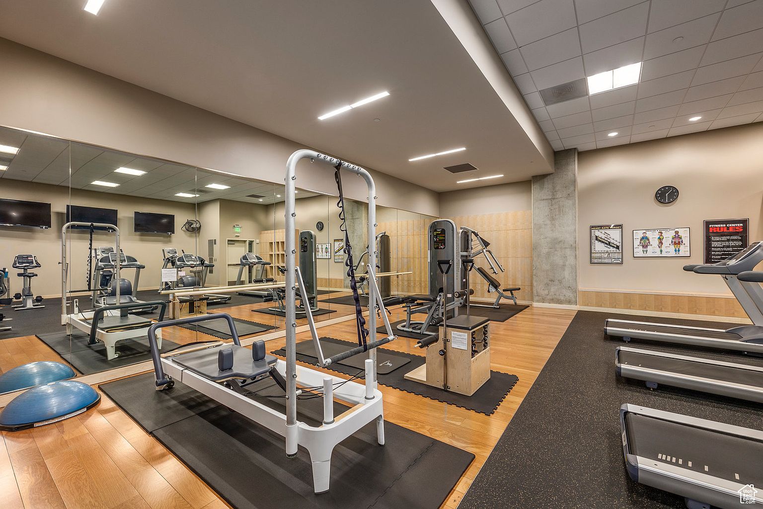 This is a well-equipped home gym featuring a variety of exercise machines including treadmills, Pilates reformers, and weight training equipment. The room has hardwood flooring in some areas and rubber flooring in others, with a large mirror spanning one wall to enhance the sense of space. The overall impression is a clean and functional fitness area.