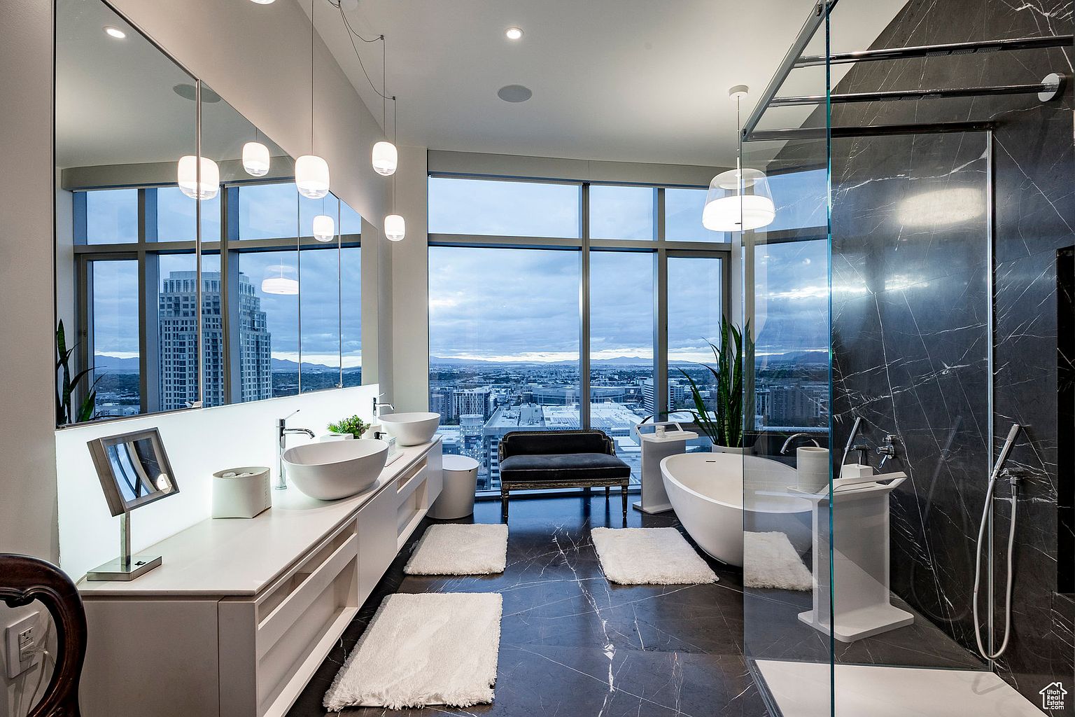 This is a luxurious primary bathroom featuring a modern design with high-end finishes. The bathroom boasts a large vanity with dual vessel sinks, a freestanding soaking tub, and a glass-enclosed shower with dark marble walls. Large windows offer stunning city views, enhancing the spa-like atmosphere.