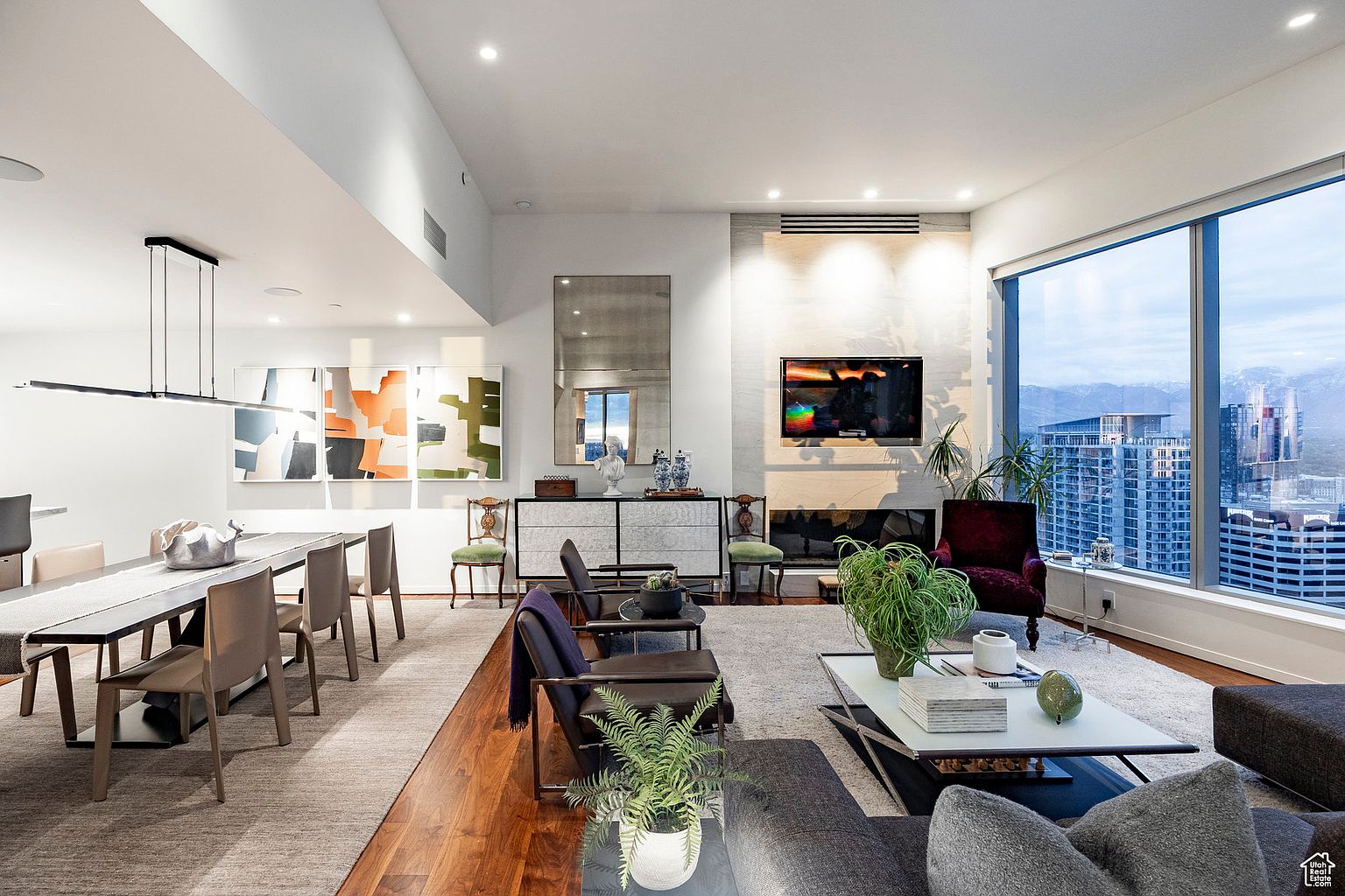 This is a spacious and modern living room with a stunning city view through a large window. The room features a fireplace with a marble surround, contemporary furniture, and stylish decor, including artwork and plants. The open layout connects to a dining area, creating a seamless and inviting living space.