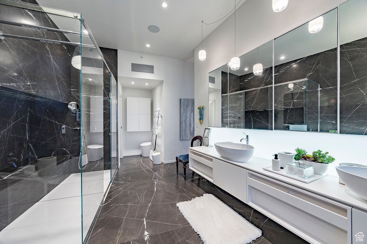 This is a luxurious primary bathroom featuring dark marble walls and flooring with white veining, creating a sophisticated and modern aesthetic. The bathroom includes a glass-enclosed shower, a double vanity with vessel sinks, and sleek white cabinetry. Pendant lights hang above the vanity, adding a touch of elegance to the space.