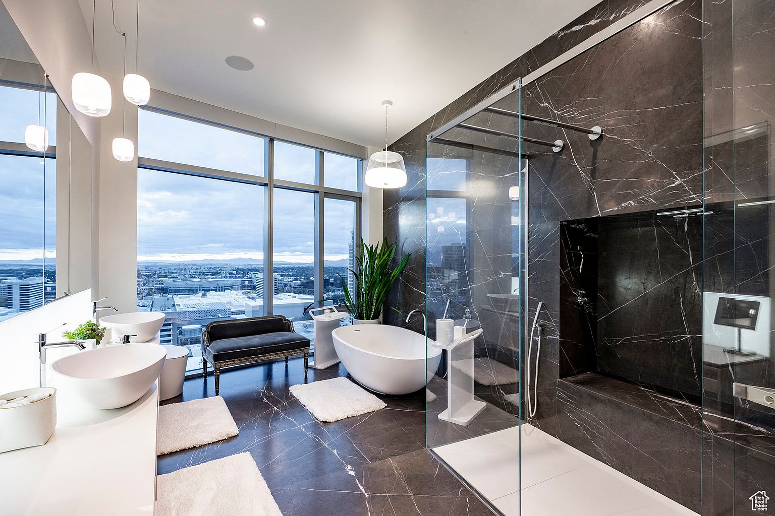 This is a luxurious primary bathroom with a modern design. It features a freestanding bathtub, dual vessel sinks on a white countertop, and a glass-enclosed shower with dark marble walls. Large windows offer a stunning city view, enhancing the spa-like atmosphere.