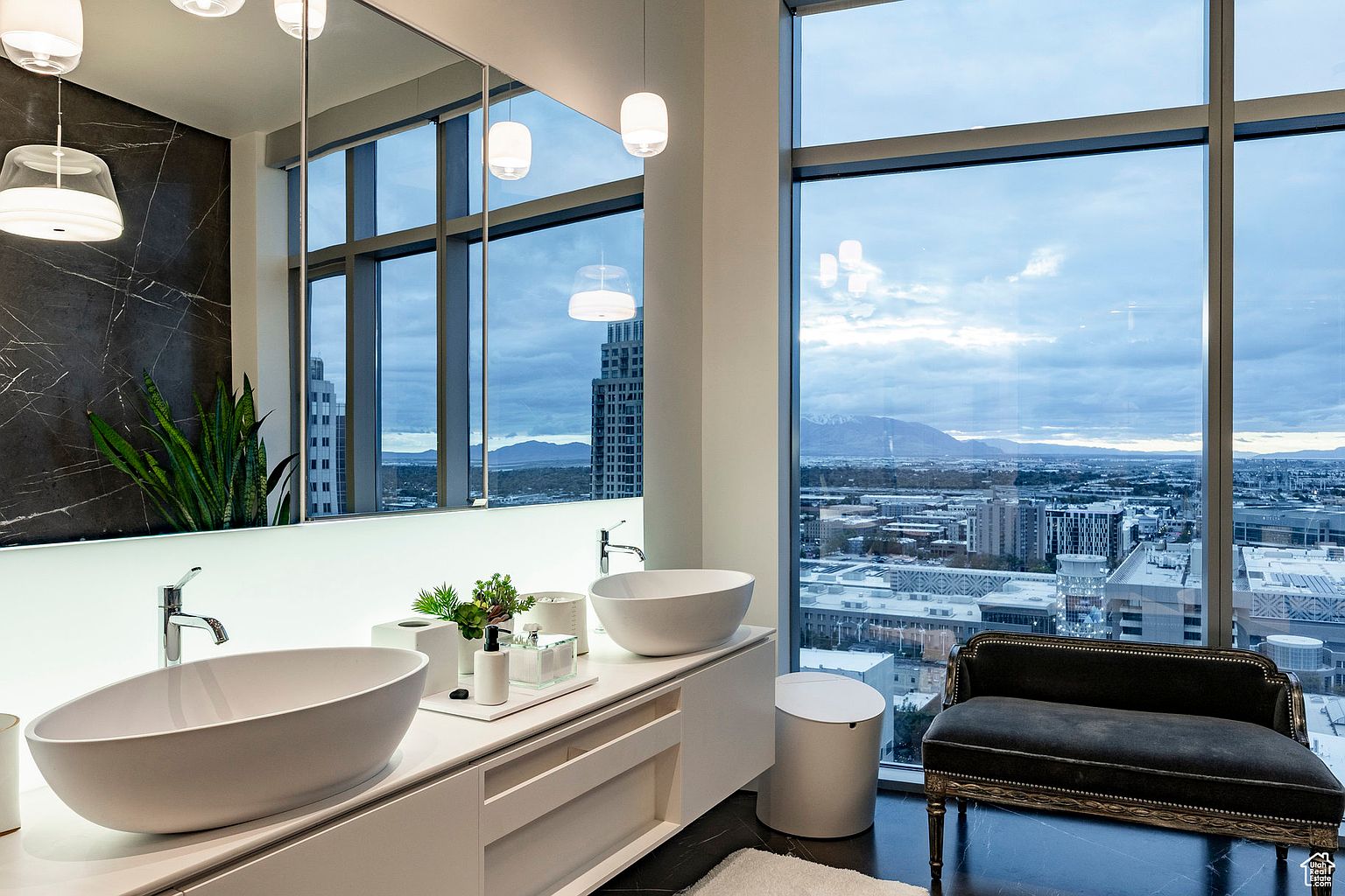 This is a luxurious primary bathroom featuring dual vessel sinks on a sleek white vanity with modern fixtures. A large mirror reflects the cityscape visible through the expansive windows, which also provide ample natural light. A stylish chaise lounge adds a touch of elegance to the space, which is finished with dark flooring and a light-colored rug.