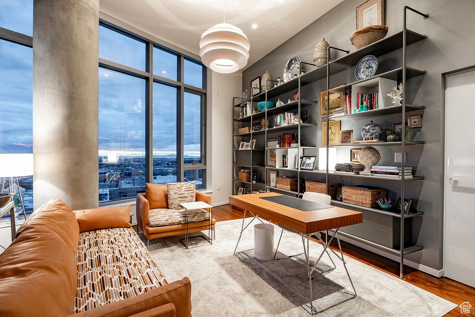 This is an interior shot of a modern office space featuring a large window with a city view, a custom shelving unit filled with books and decorative items, and a sleek wooden desk with metal legs. The room is furnished with a comfortable sofa and chair, creating a stylish and functional workspace. The overall aesthetic is sophisticated and inviting.