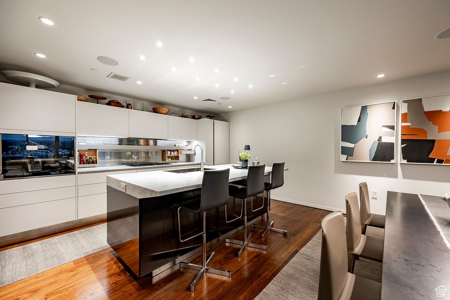This is a modern kitchen with sleek white cabinetry and a dark island with a white countertop. Three bar stools are positioned at the island, and there is a dining area with chairs visible in the foreground. The kitchen features stainless steel appliances and a minimalist design, creating a clean and sophisticated atmosphere.